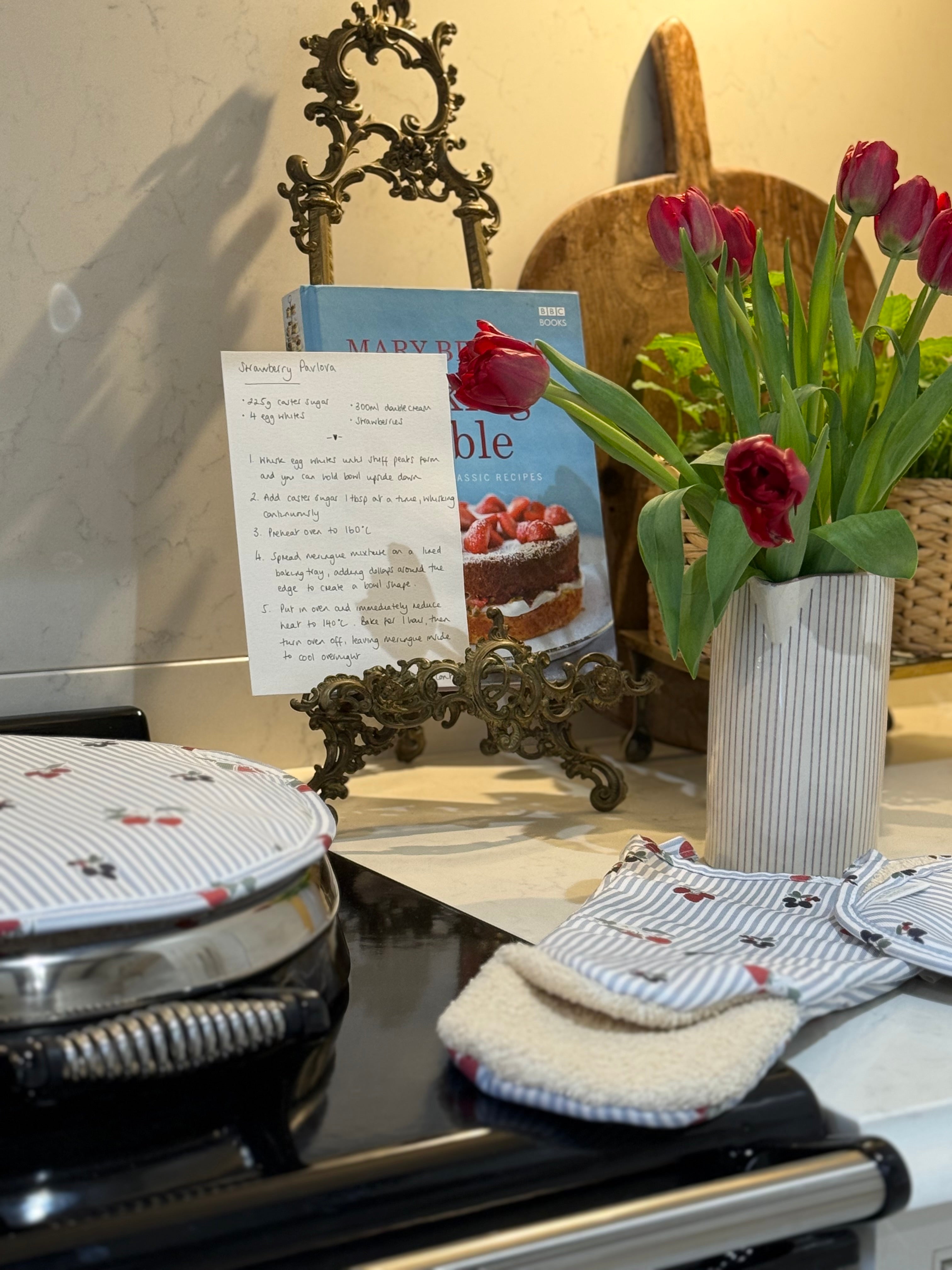 Blue & white striped oven gauntlet with berry design on a range cooker with flowers and recipes in background