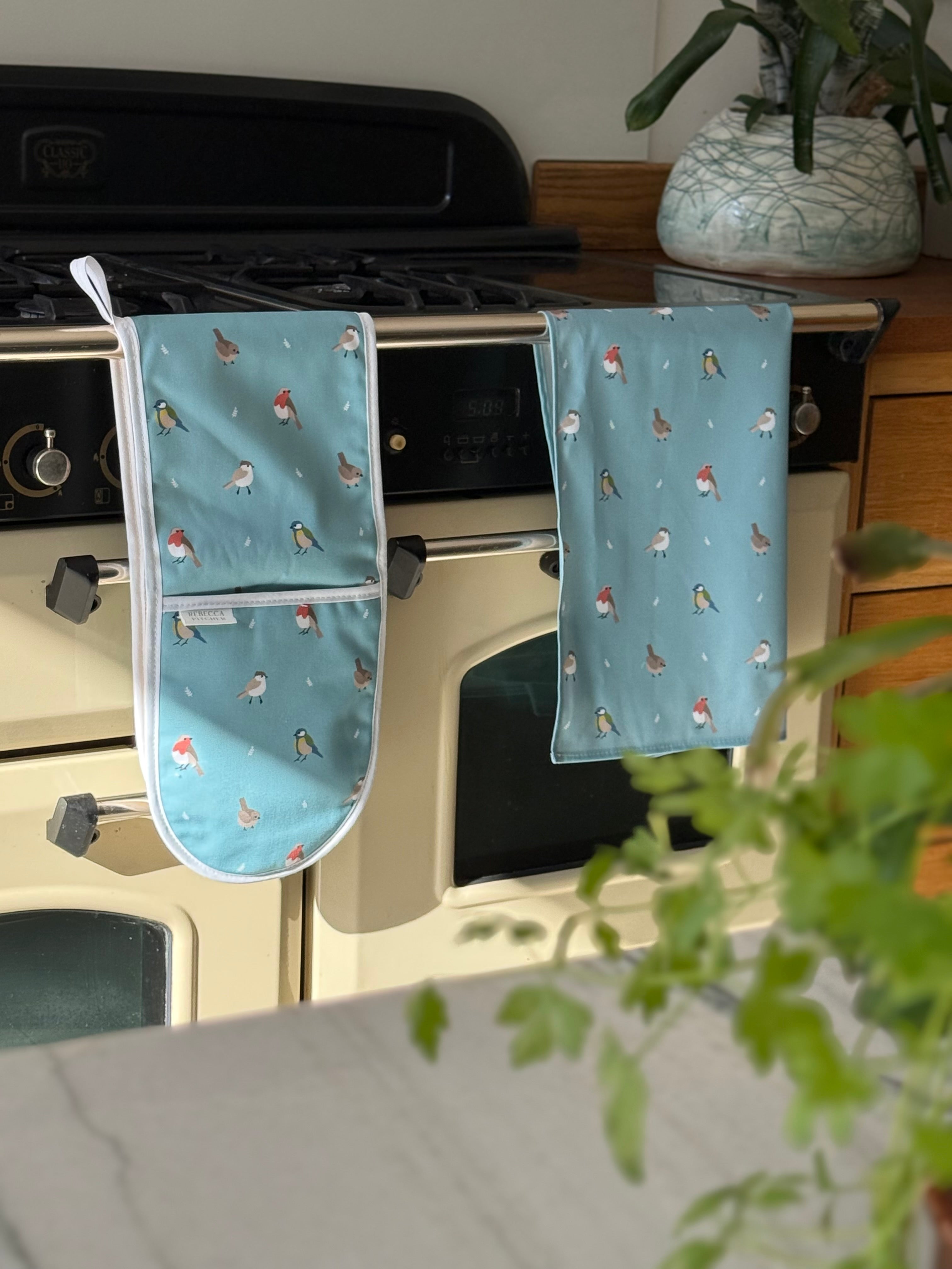 Pair of blue oven mitts with bird pattern hanging on a stove.