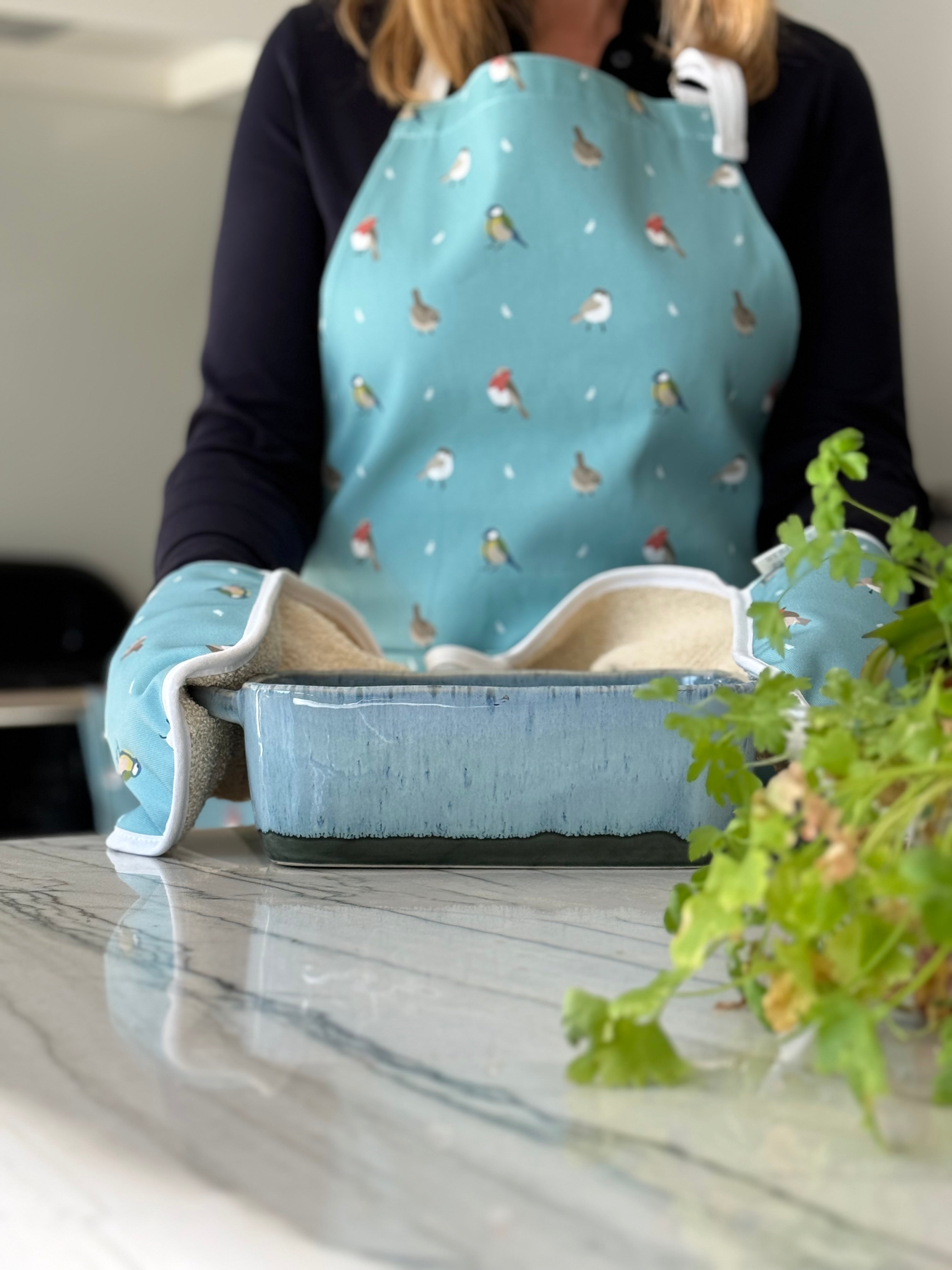 Person wearing a blue apron with bird pattern with a cooking dish on a marble kitchen worktop