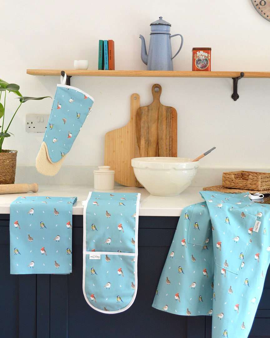 Kitchen baking setting with ocean blue bird patterned oven mitt, gauntlet, apron and towel on a white countertop.