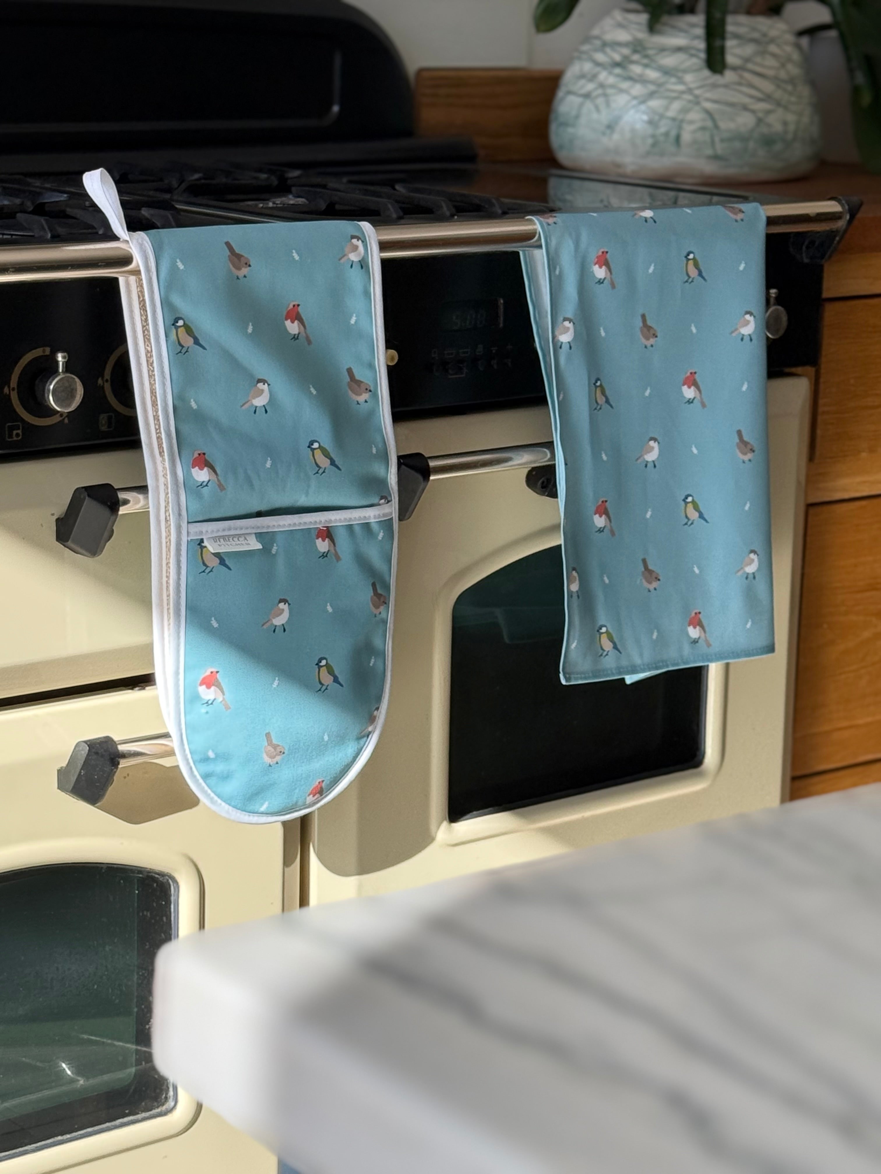 Blue tea towel and oven mitts with bird pattern hanging on a stove.