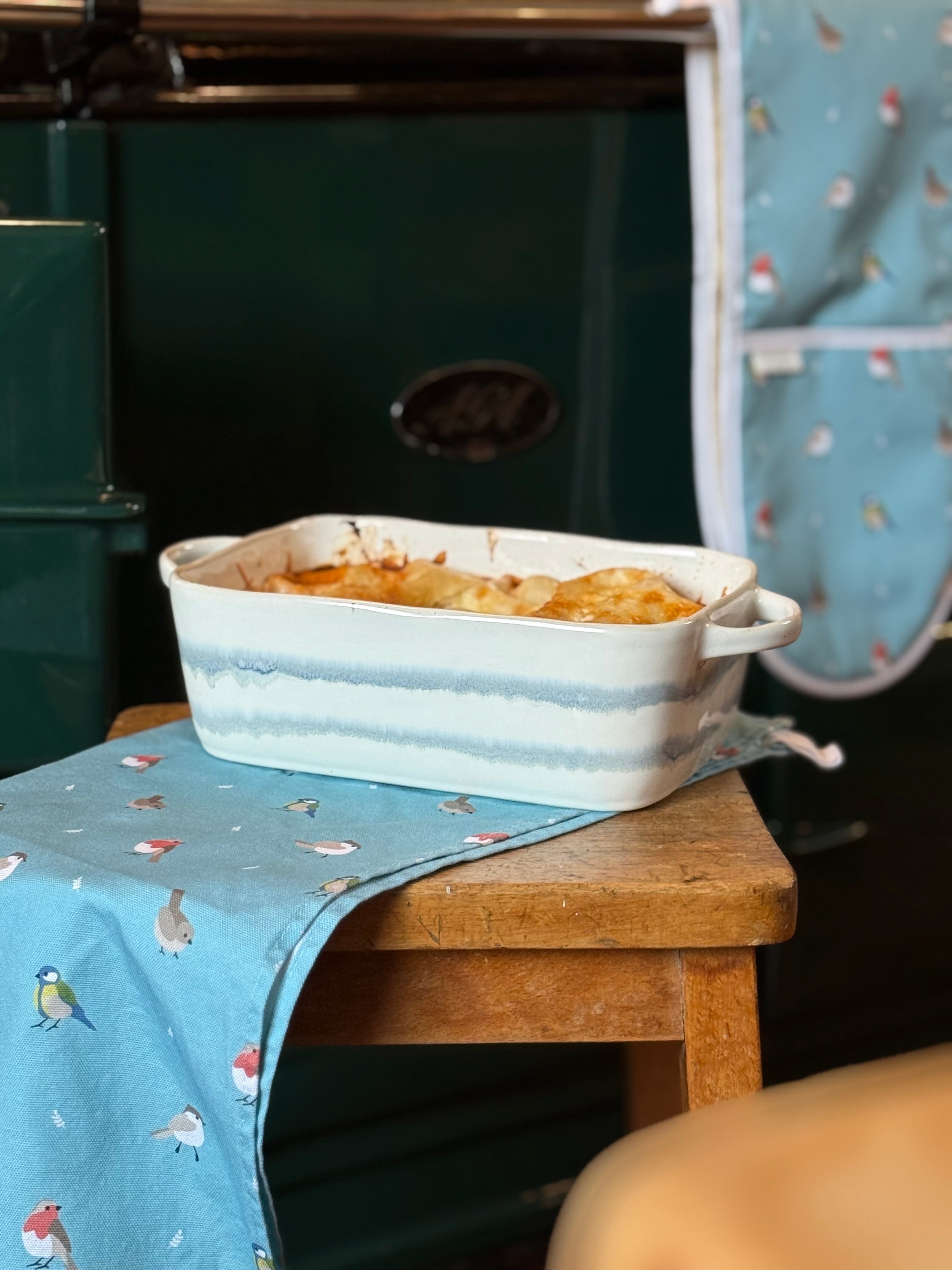 Casserole dish with food on a wooden stool with bird patternered kitchen textiles in front of an range cooker.