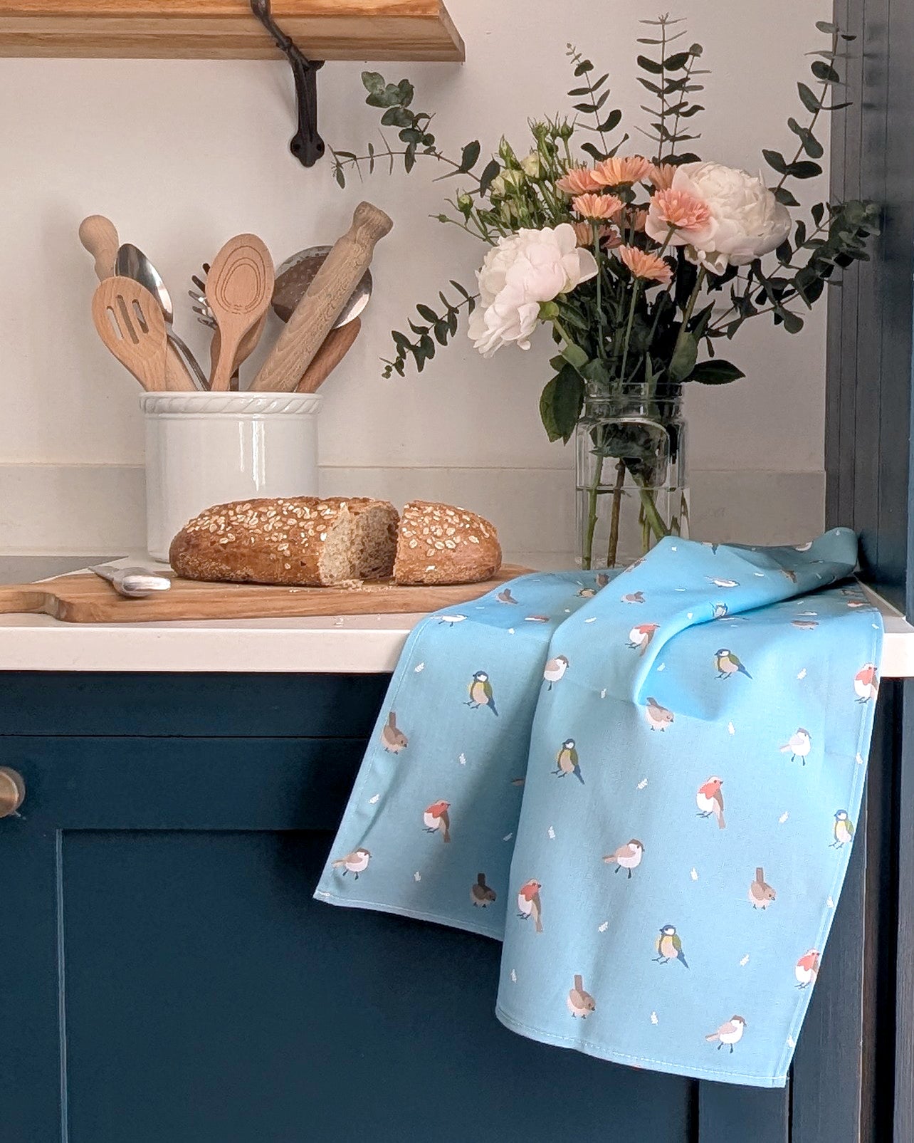 Kitchen scene with bread on a cutting board, flowers in a vase, and a blue towel with bird pattern.