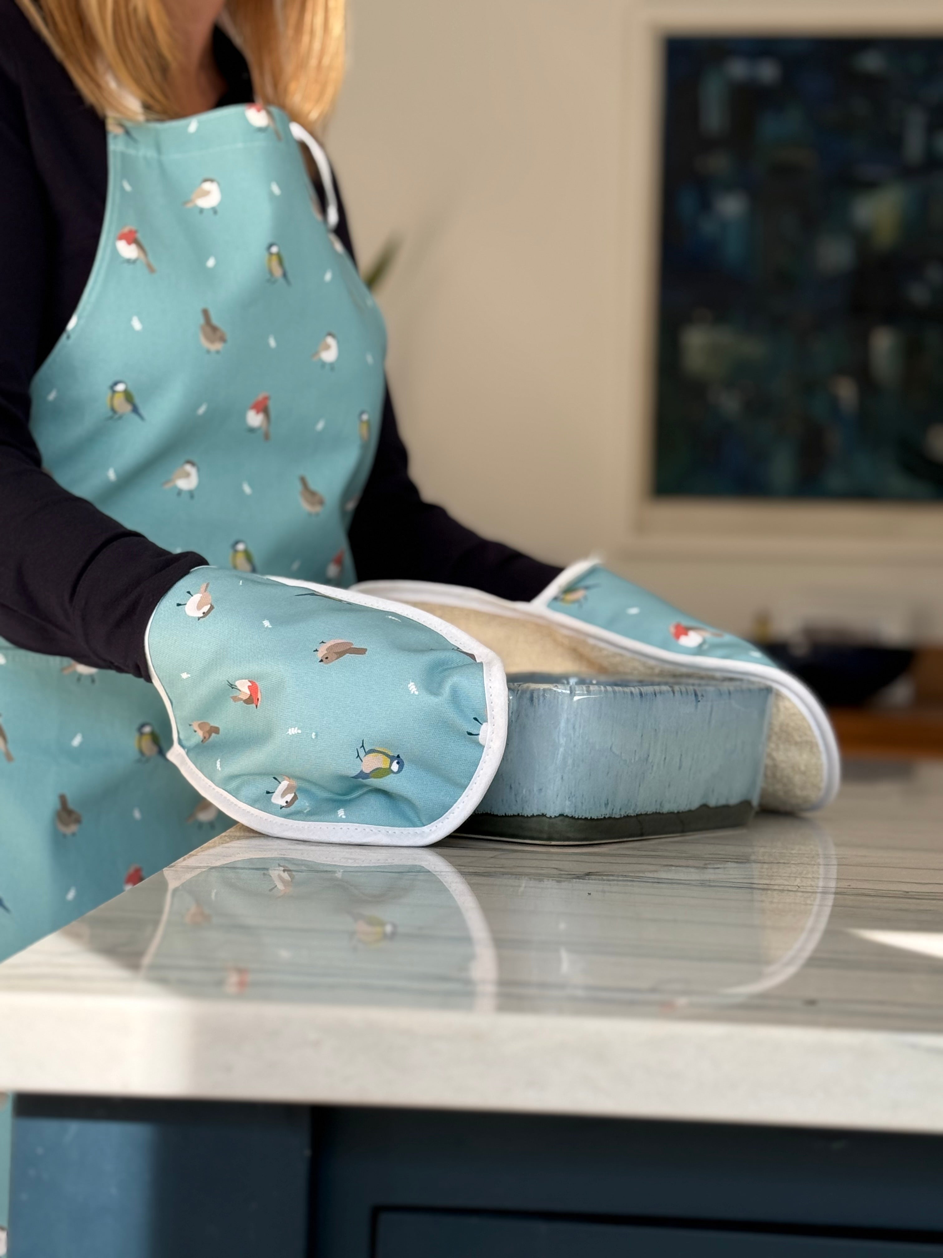 Person cooking with bird pattern oven gloves wearing an apron in a kitchen setting