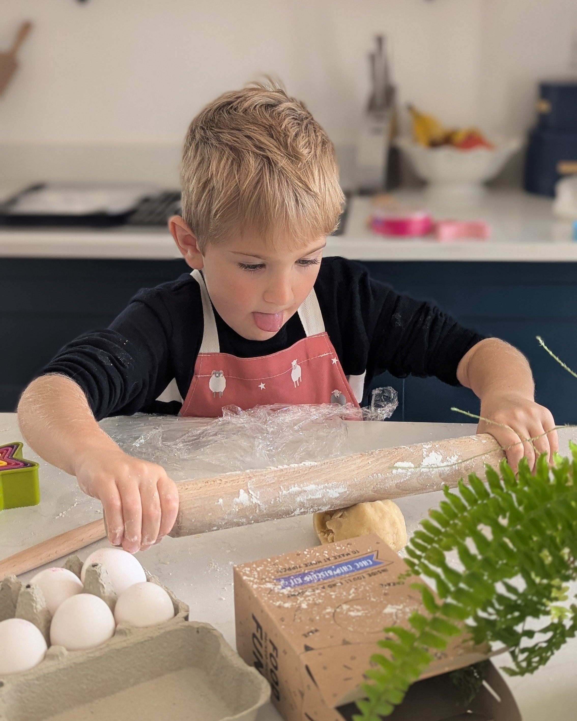 Child wearing red sheep design apron in a kitchen using a rolling pin on a counter with eggs and a box in the foreground.