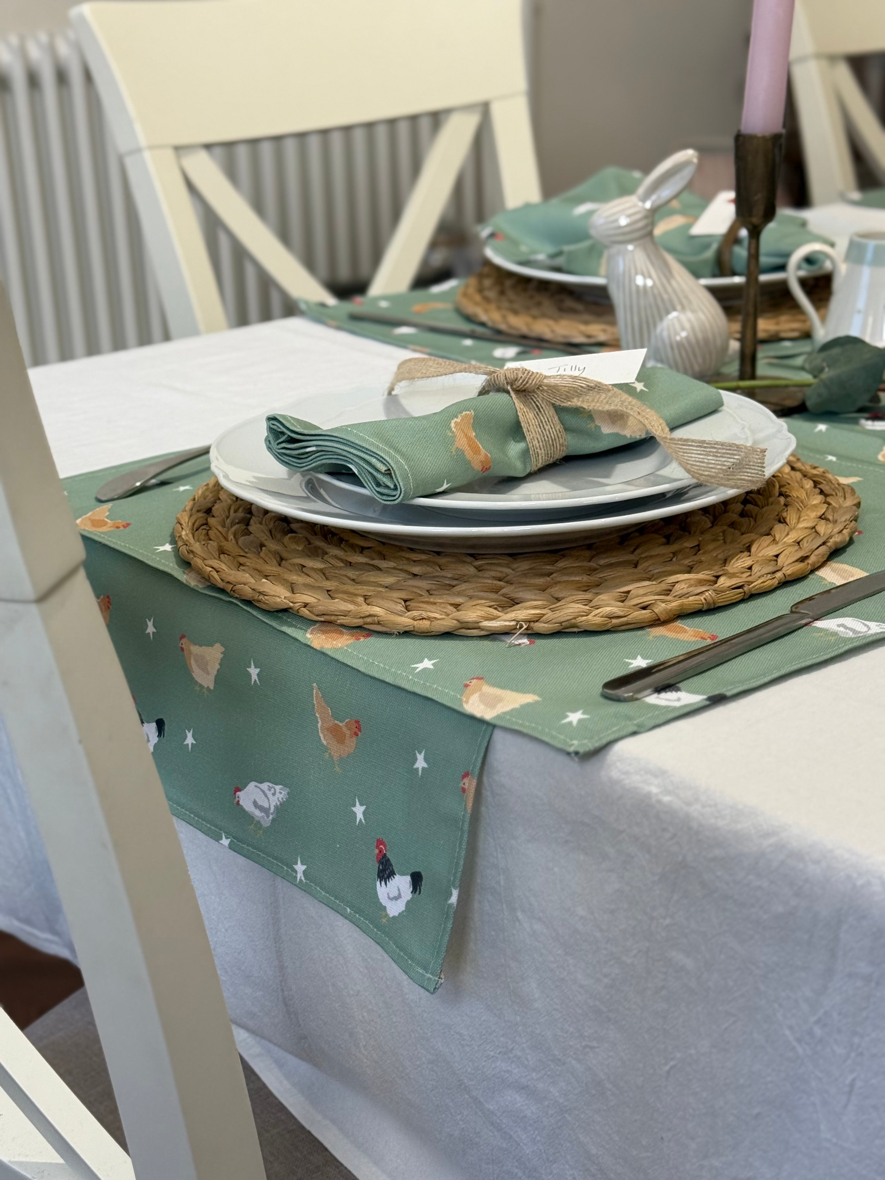 Dining table setting with a green table runner, placemats and napkins featuring chicken designs.