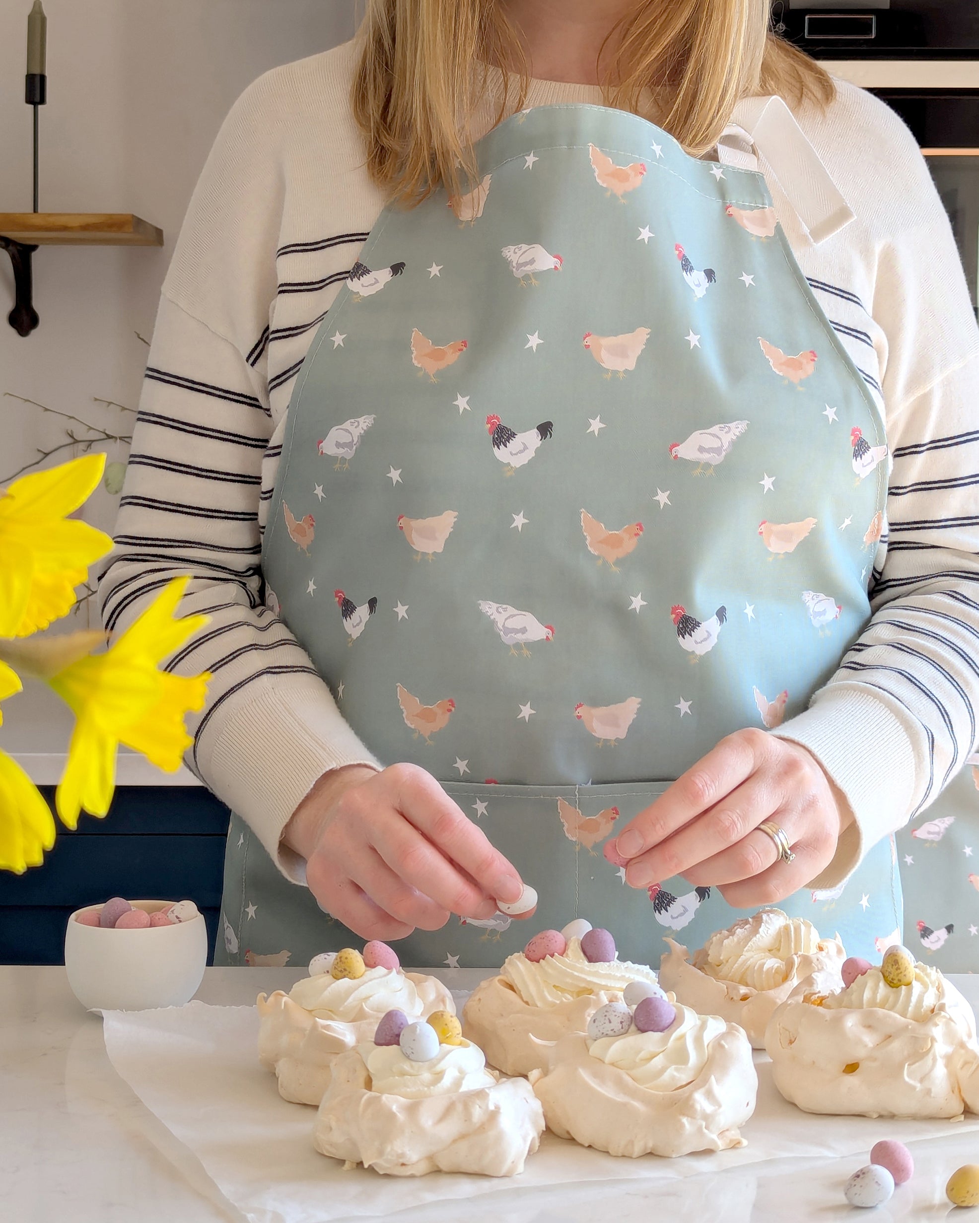Rebecca Pitcher in her kitchen wearing a chicken patterned apron, preparing dessert with meringues and colorful eggs.