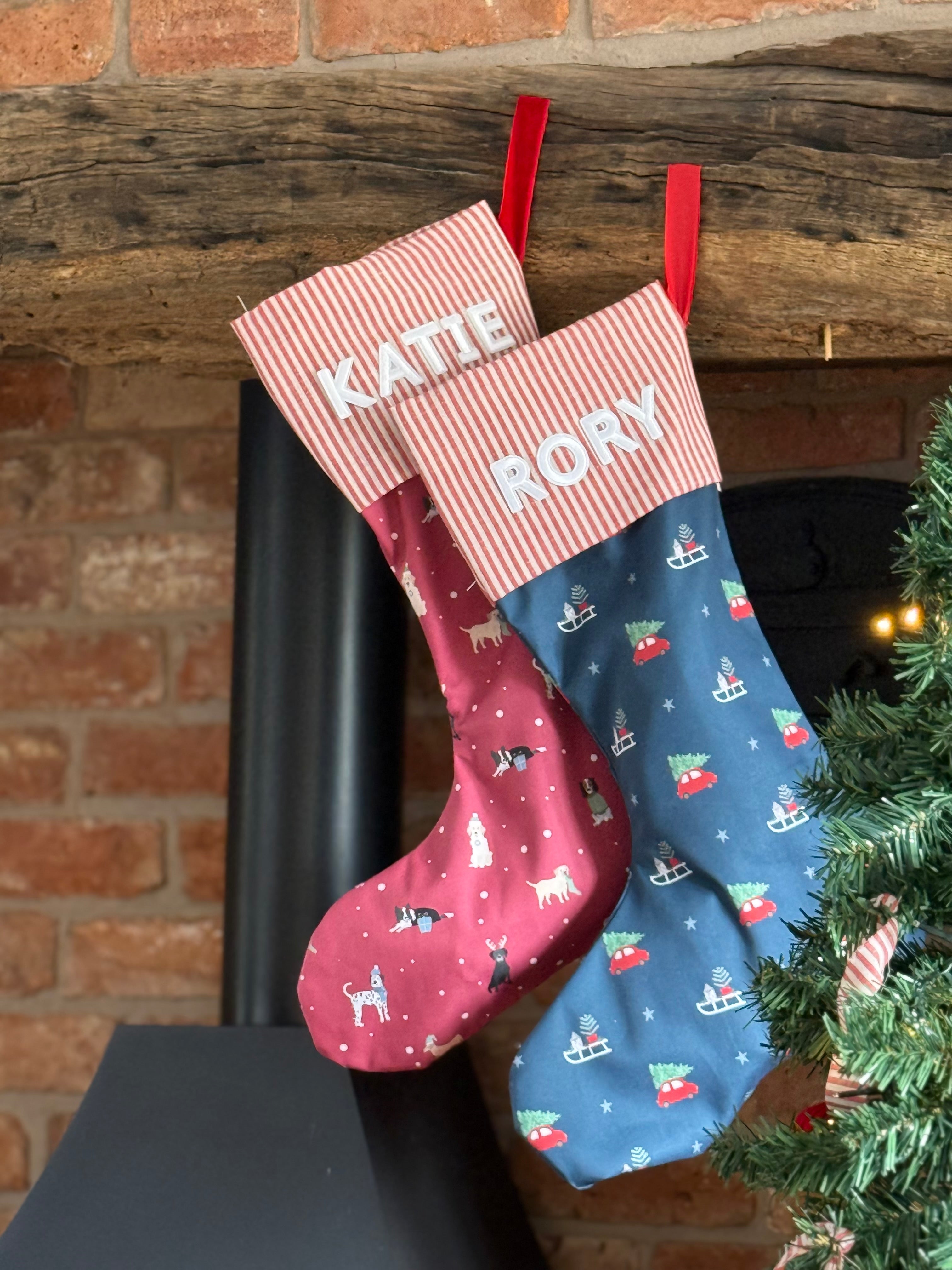 Two Christmas stockings, one red with dog pattern and one blue with Christmas trees and cars, hanging on a fireplace with a tree in the background.