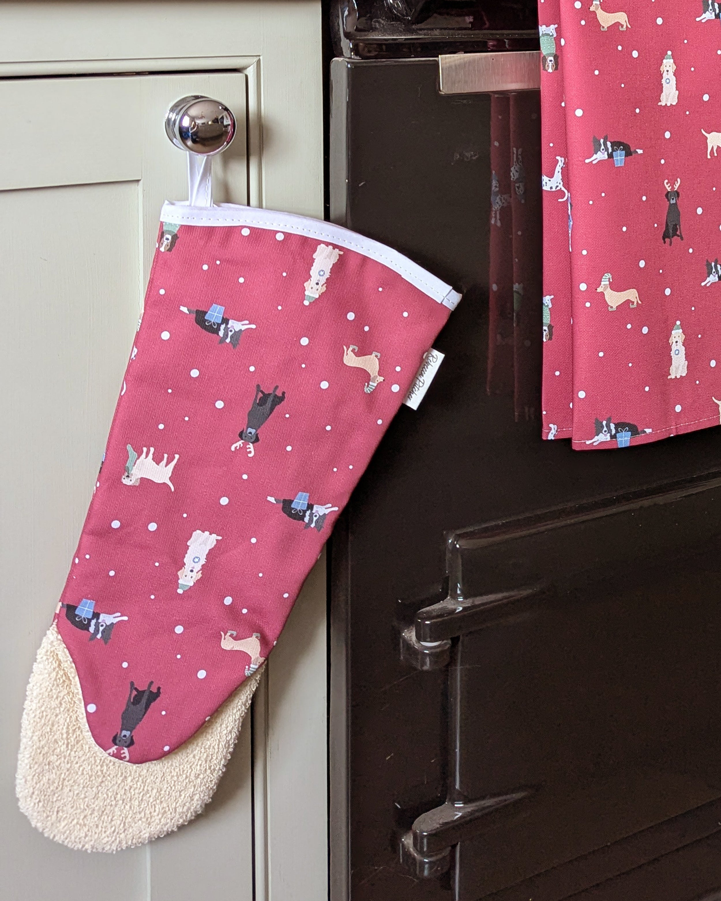 Red long oven mitt and tea towel with Christmas dogs pattern hanging on a kitchen cabinet.