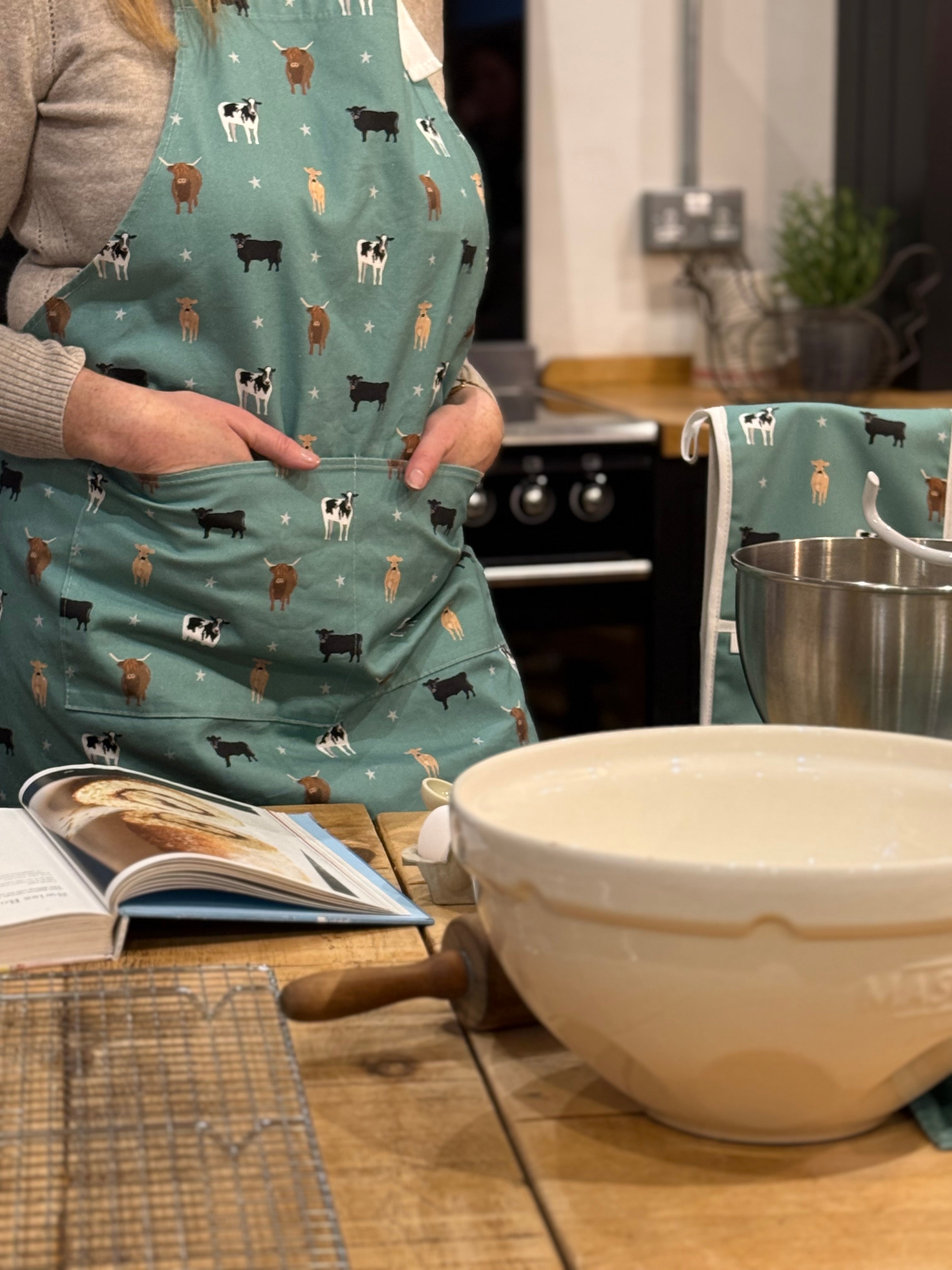 Person wearing a cow-patterned apron in a kitchen with a bowl and rolling pin on the counter.