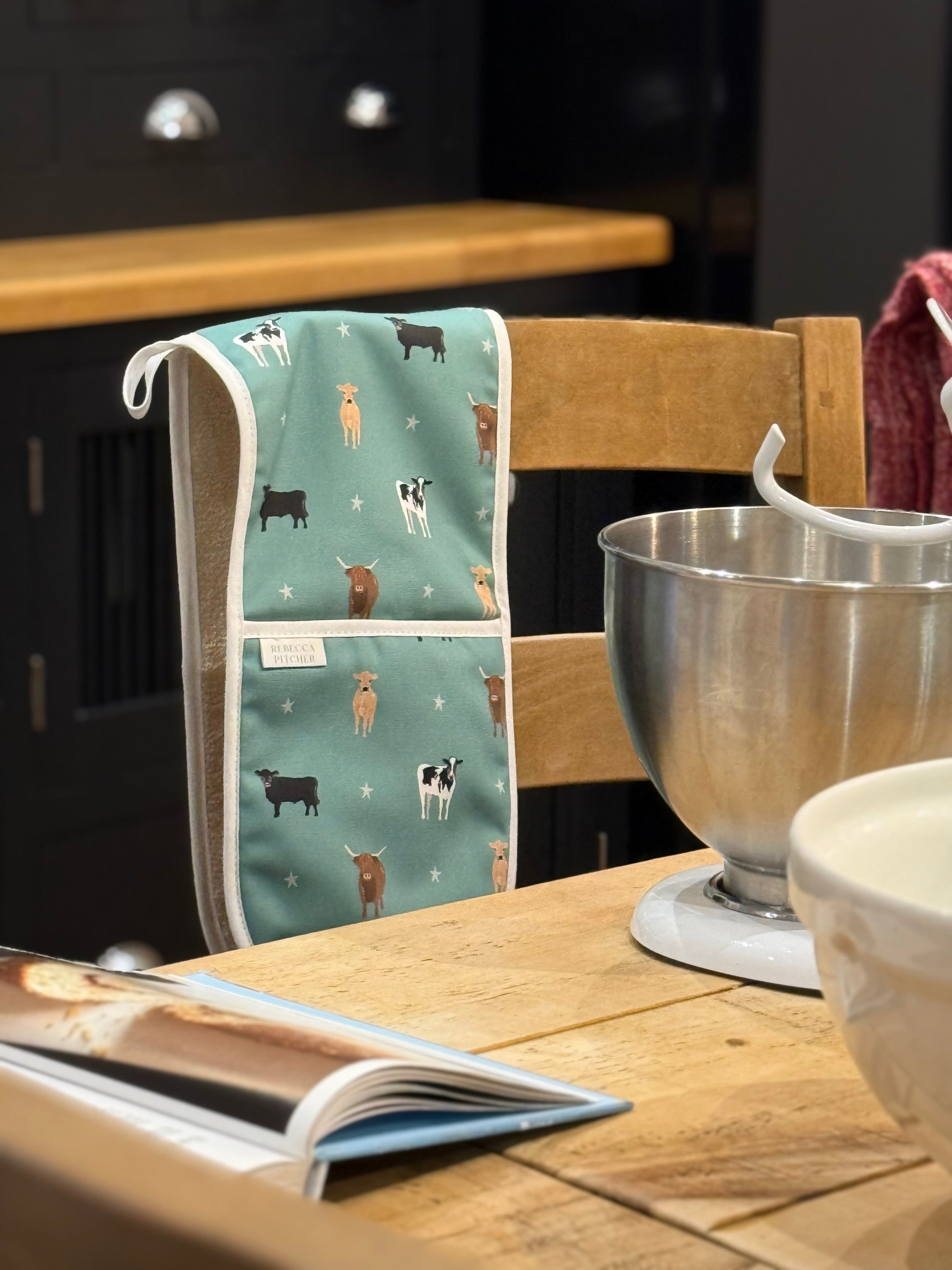 Kitchen scene with a cow patterned double oven glove hanging on a chair, mixing bowl, and cookbook on a table.