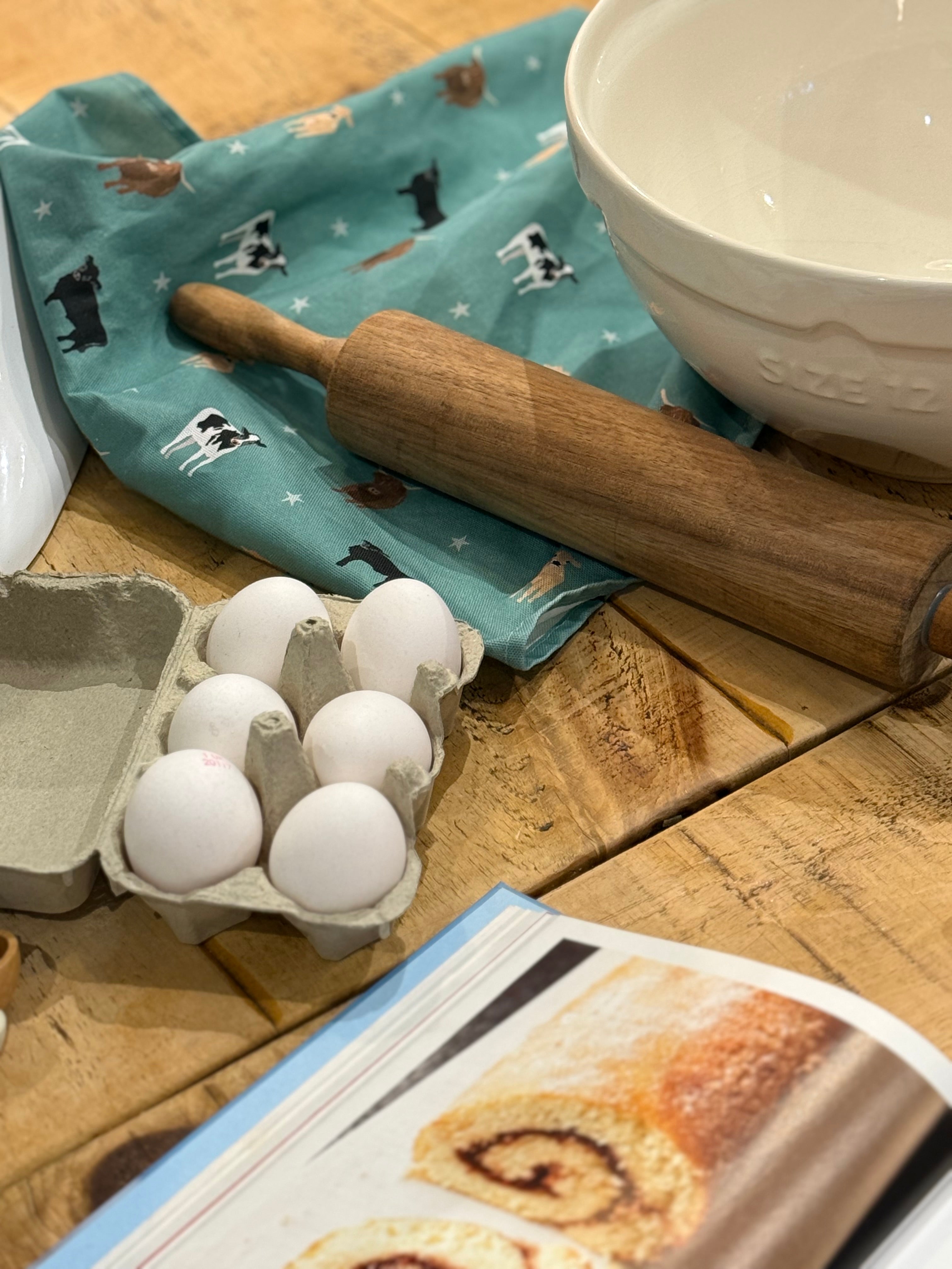 Wooden rolling pin, eggs in a carton, and a cookbook on a wooden table with a green cow design tea towel.