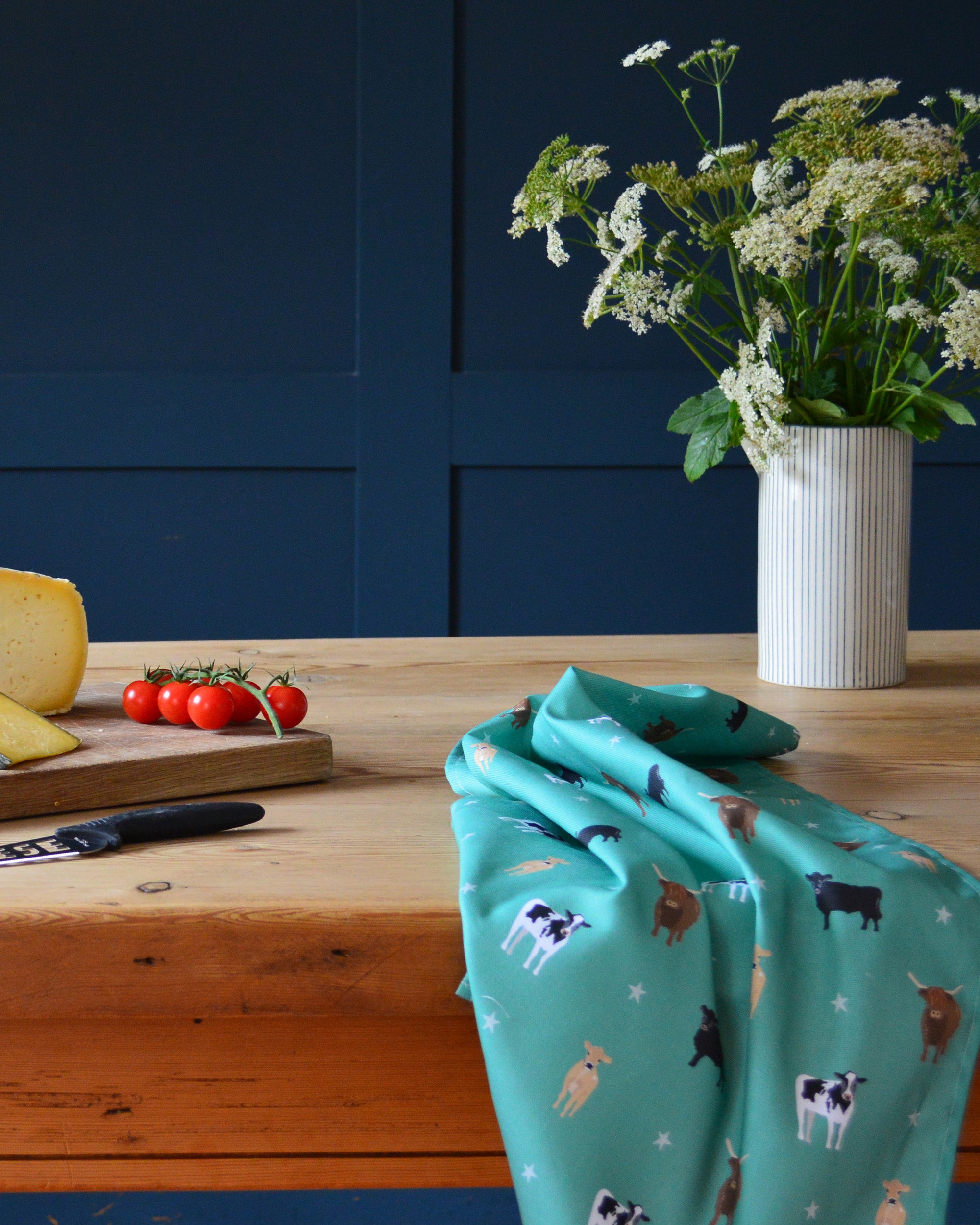 Green cotton tea towel with cow pattern on a wooden table against a blue wall.