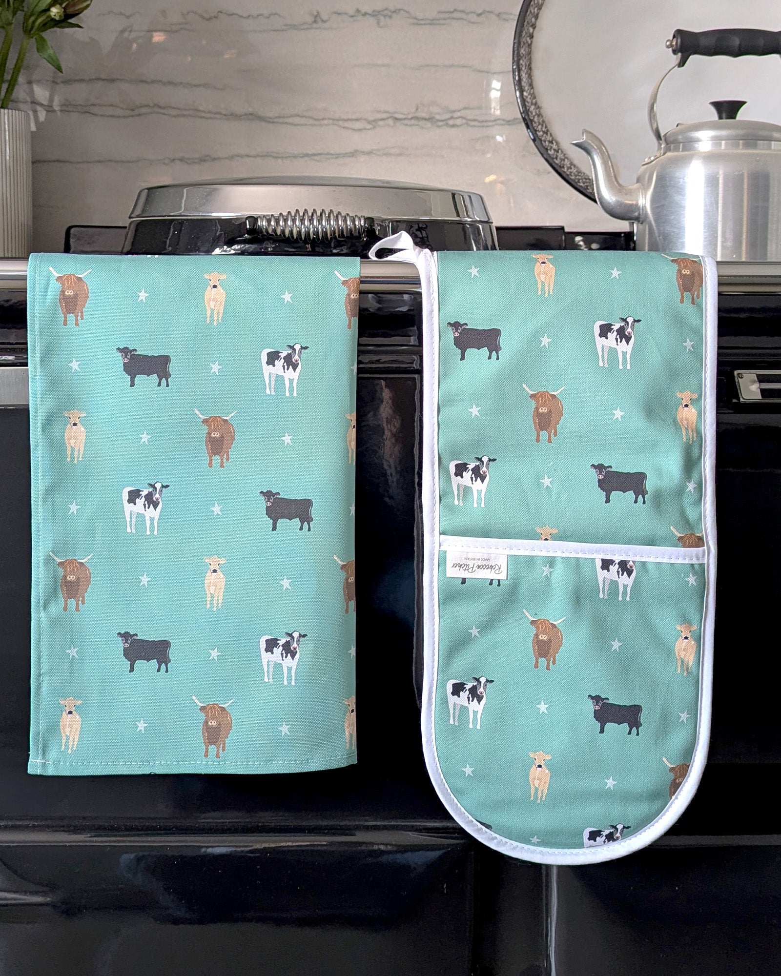 Green tea towel and oven mitt with cow pattern hanging on a stove.