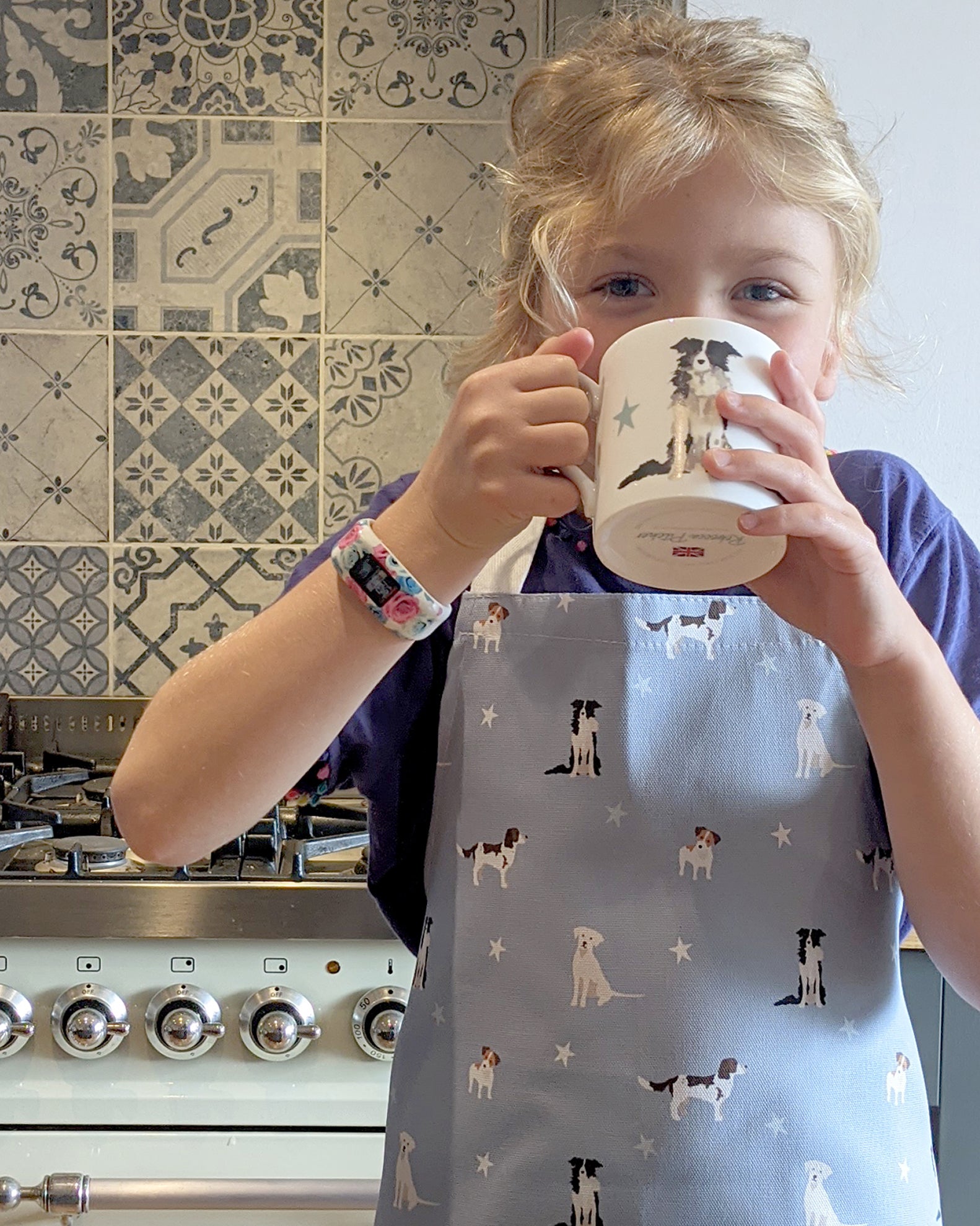 Child in a kitchen wearing a dog-themed apron and holding a mug.
