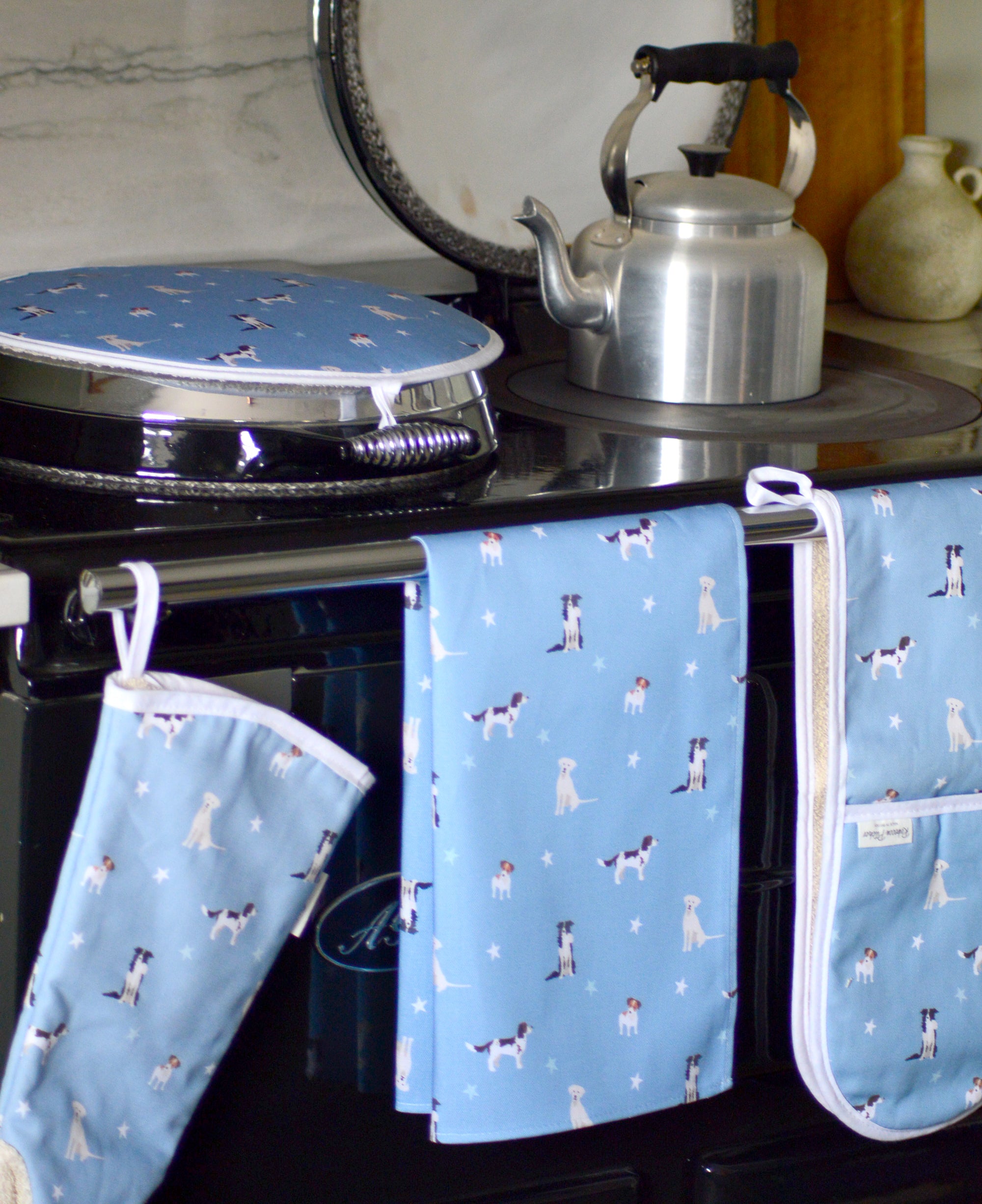 Blue tea towel with dog pattern hanging on a stove with a kettle oven mitts and hob cover