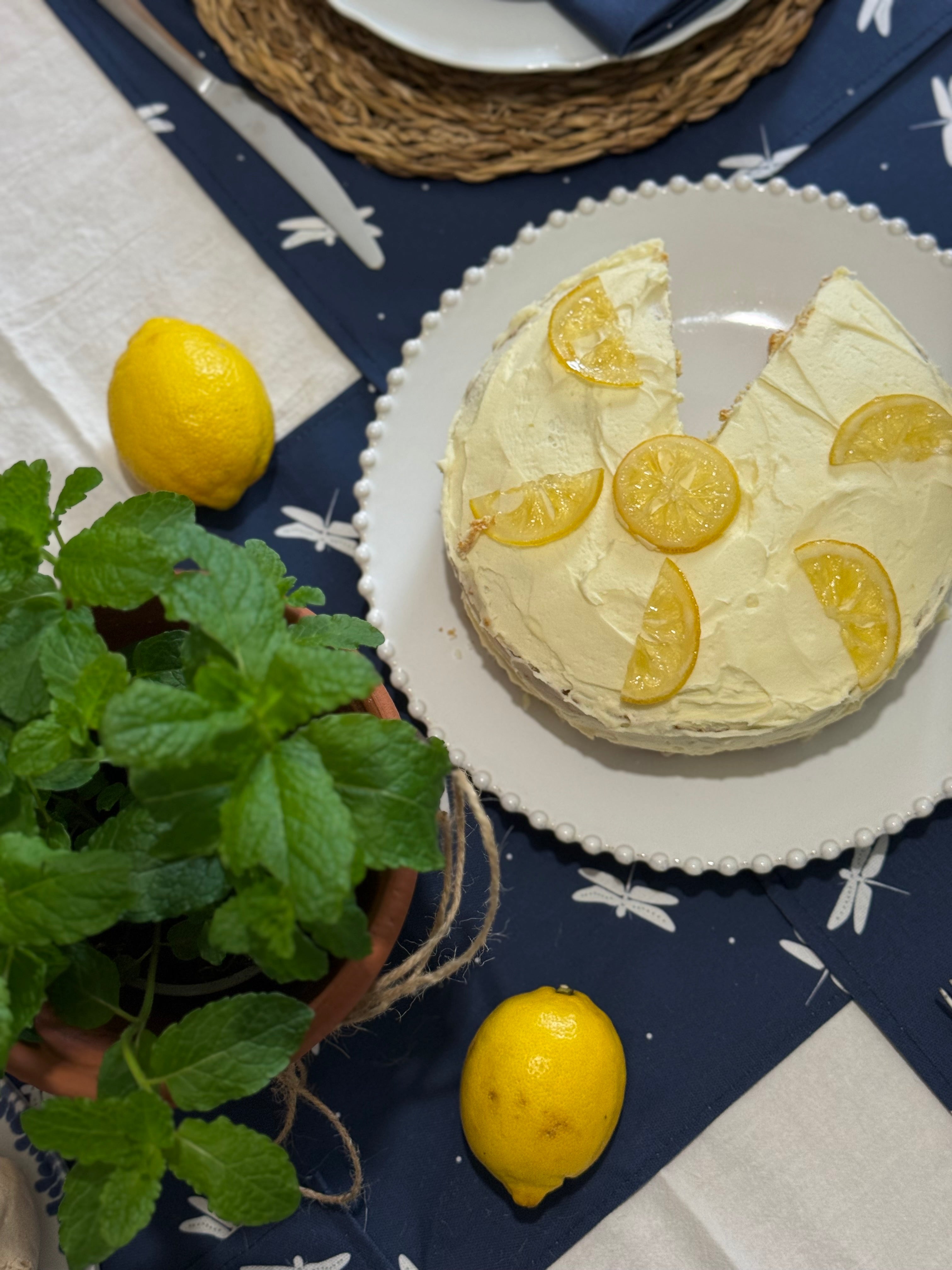 Lemon cake with a slice taken out on a navy dragonfly table runner with lemons and a plant.