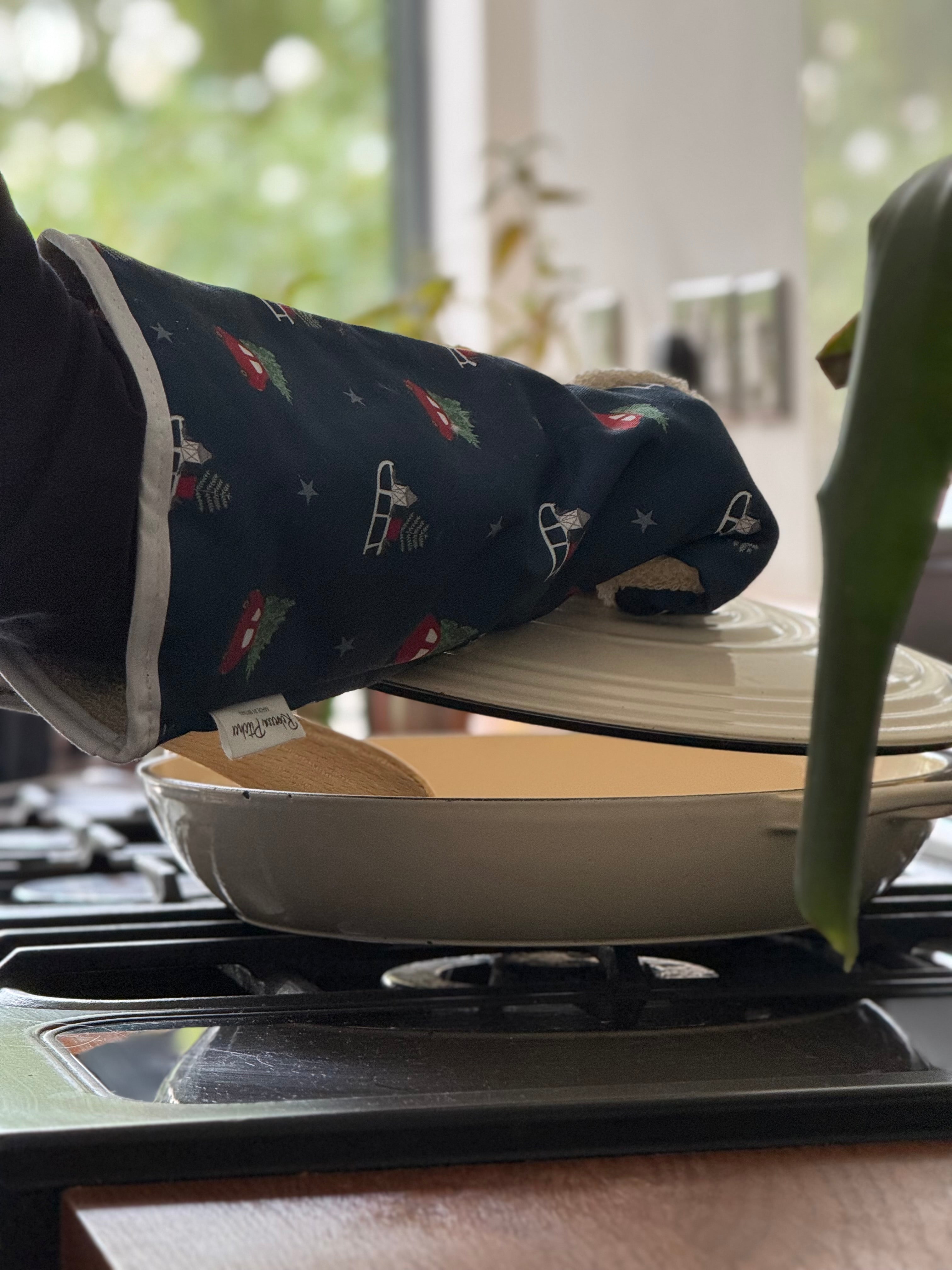 Person wearing a navy blue oven mitt with Driving Home for Christmas design holding a white pot on a stove.