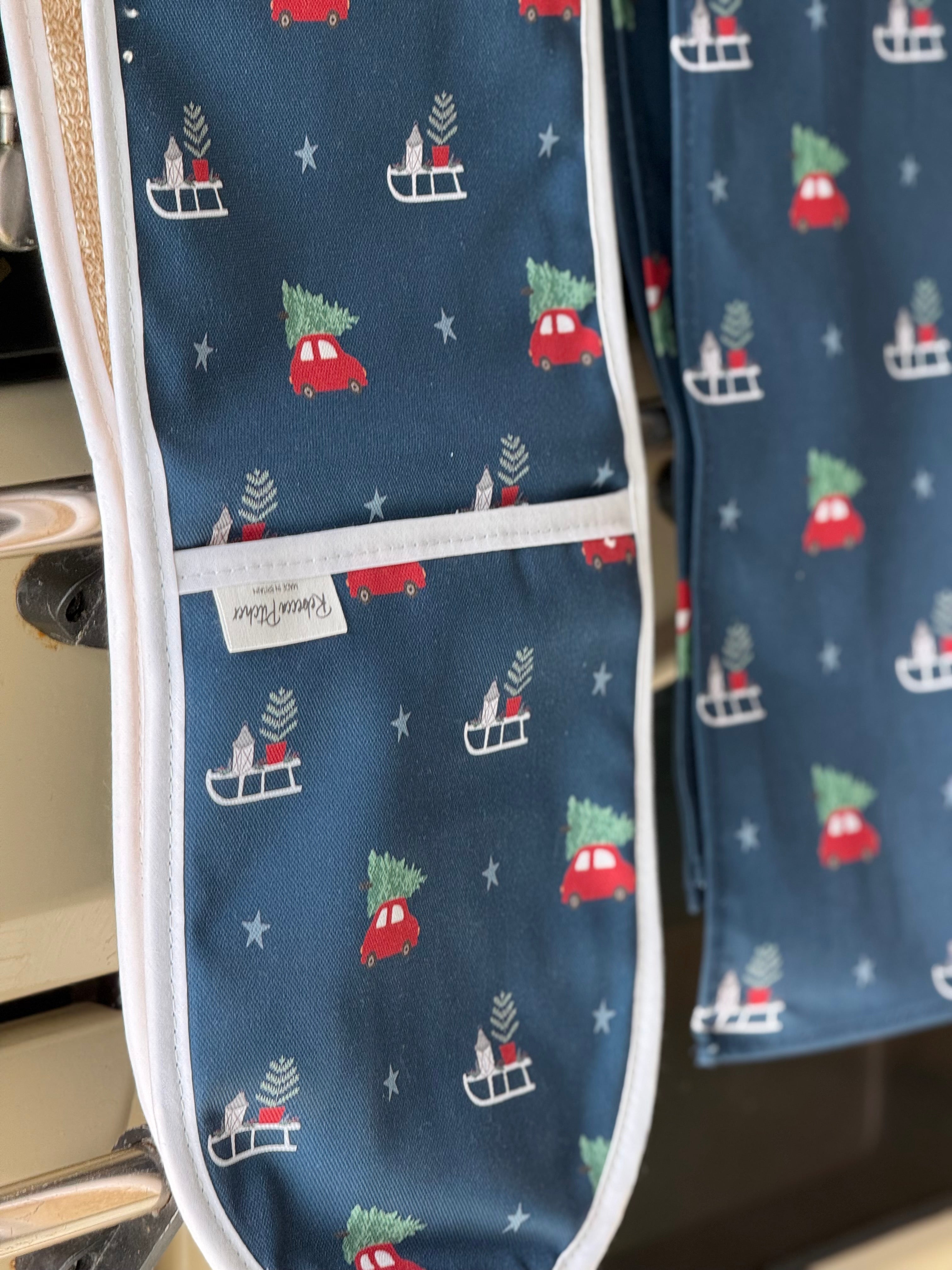 Blue double ovengloves with Christmas tree and car pattern hanging on a cooker rail.