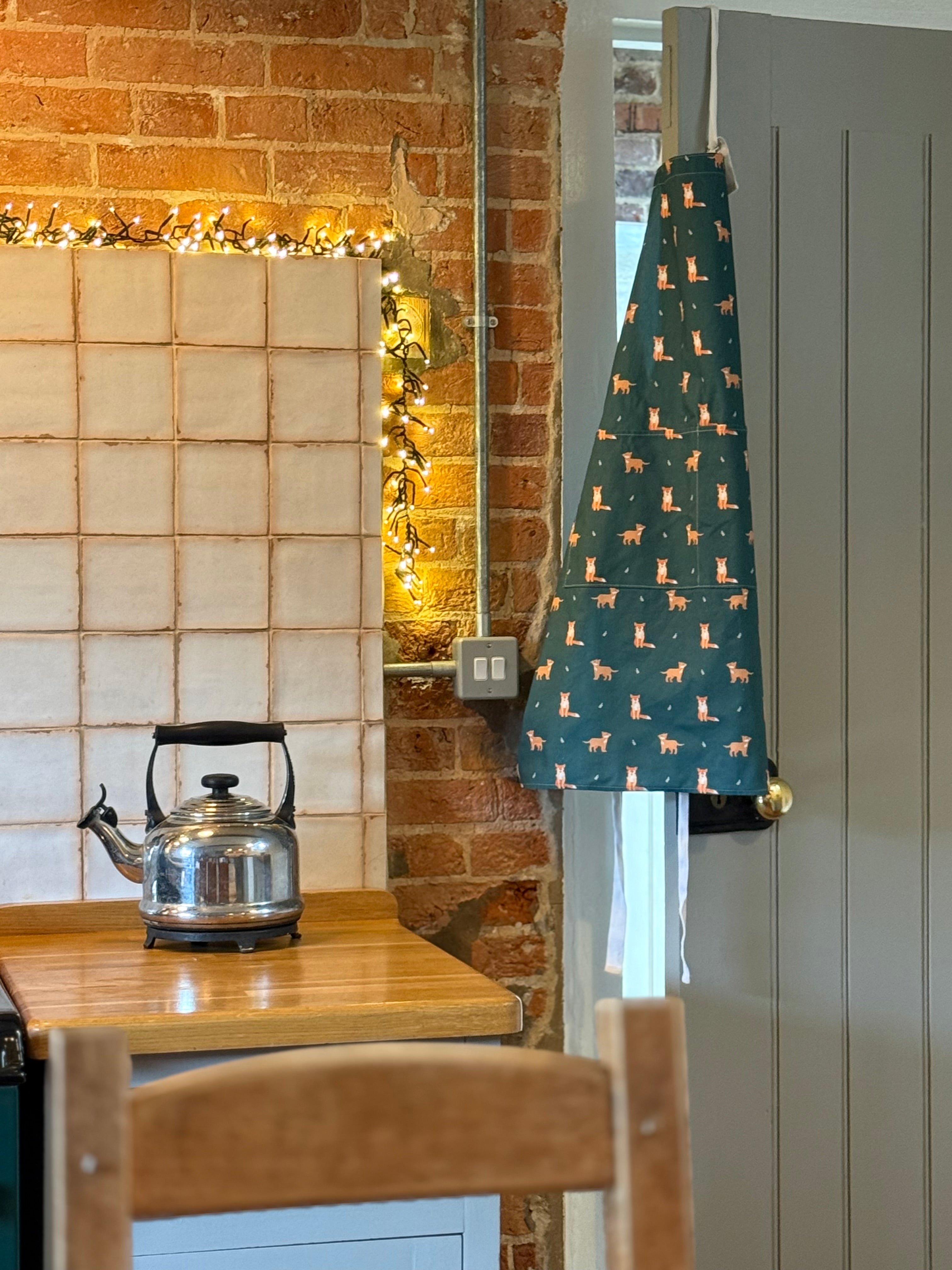 Kitchen interior with a kettle on a wooden counter, decorative lights on the wall, and a green fox patterned apron hanging on a door