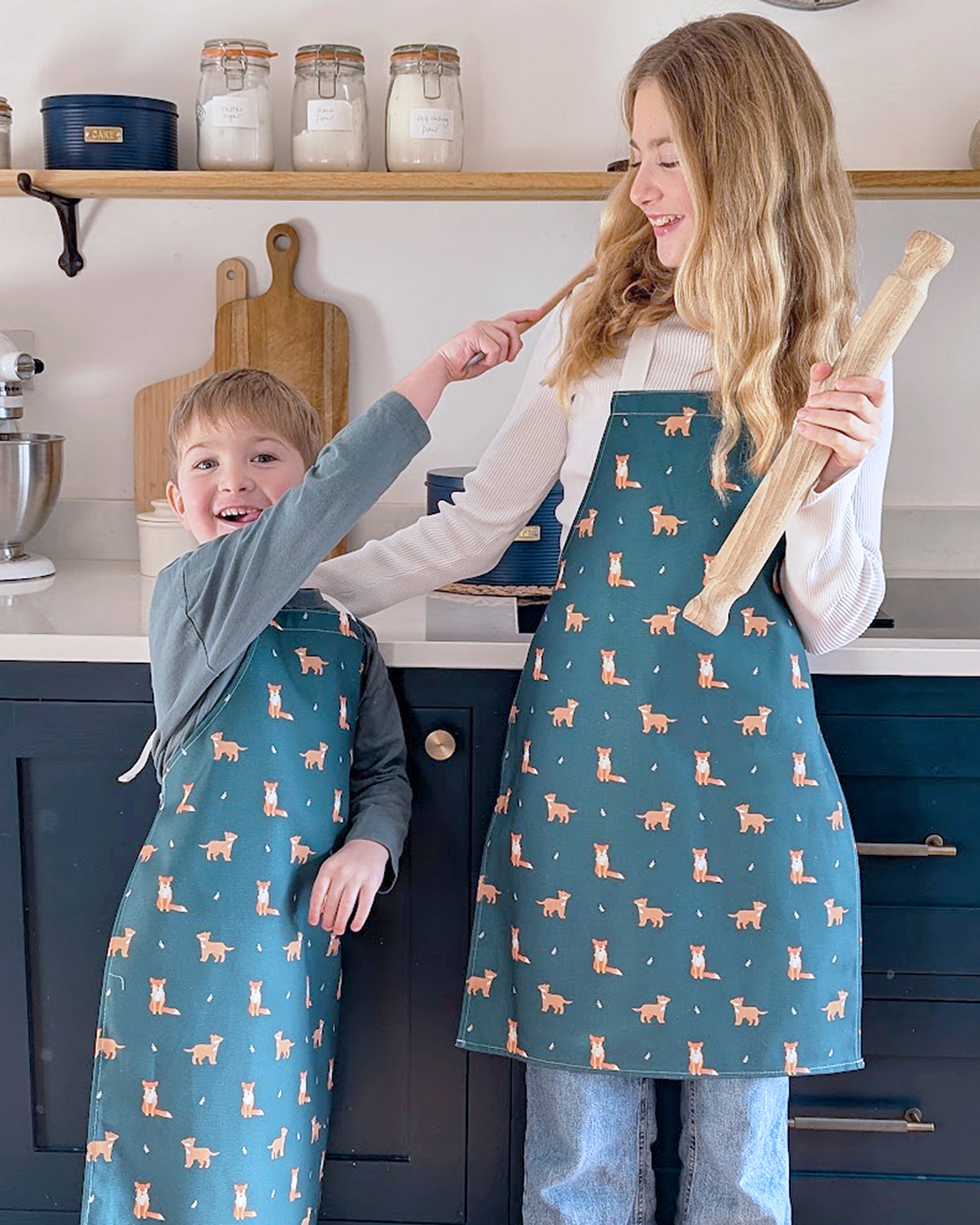 Two children in a kitchen wearing green aprons with fox patterns, one holding a rolling pin.