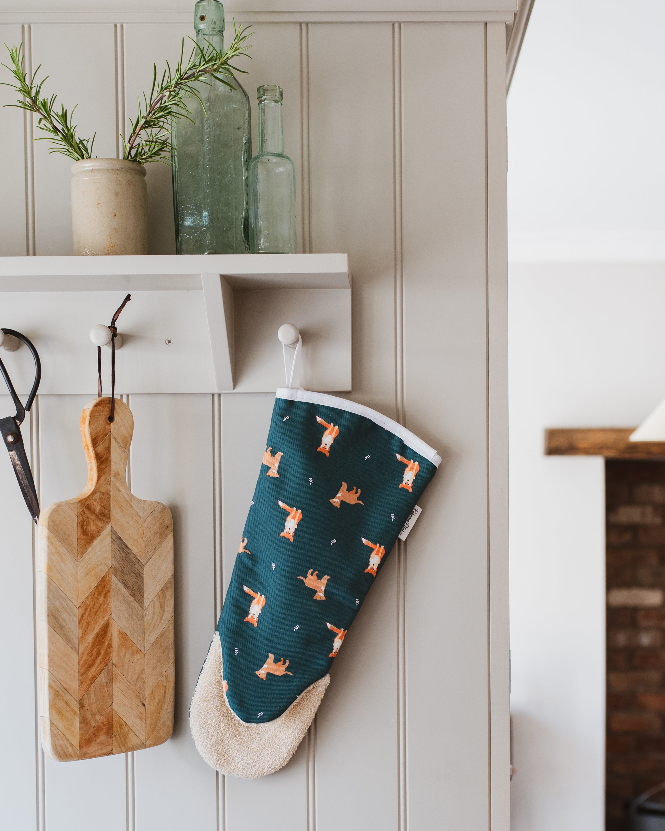Oven mitt with fox pattern hanging on a white wall next to a wooden cutting board.
