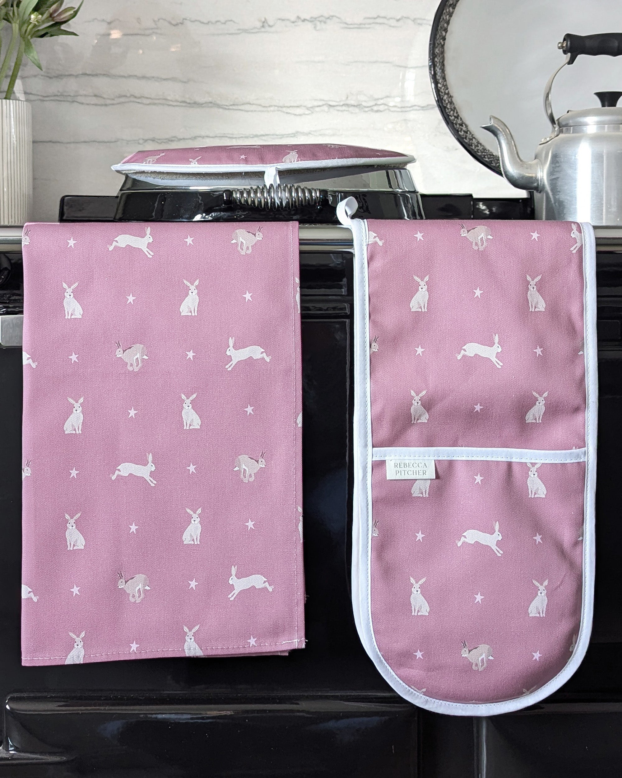 Pink oven mitt, tea towel and hob cover with hare pattern on a stove.