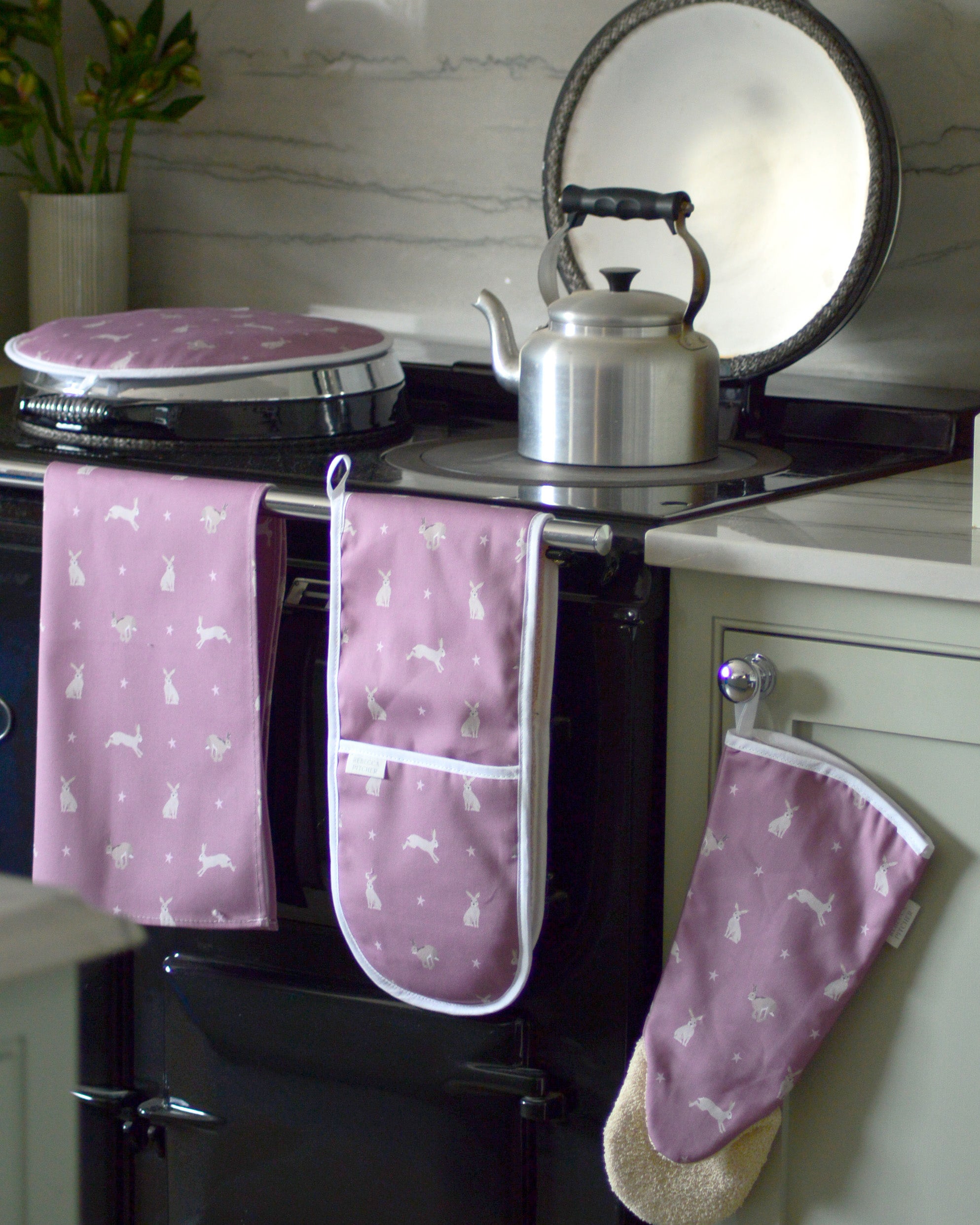 Pink oven mitts with hare pattern hanging on a black stove.