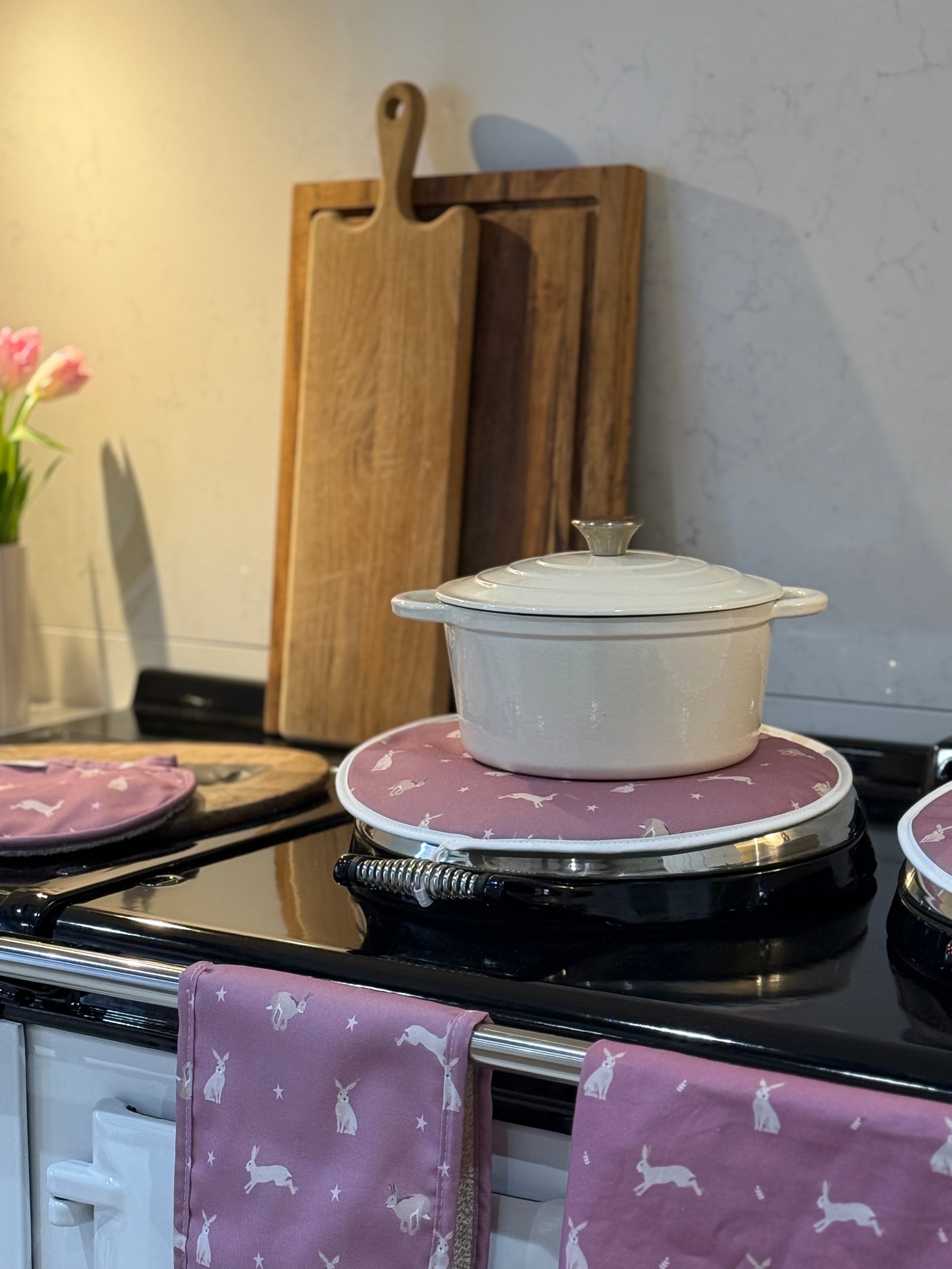 White casserole dish on a stove with pink hare design hob cover and cutting boards in the background