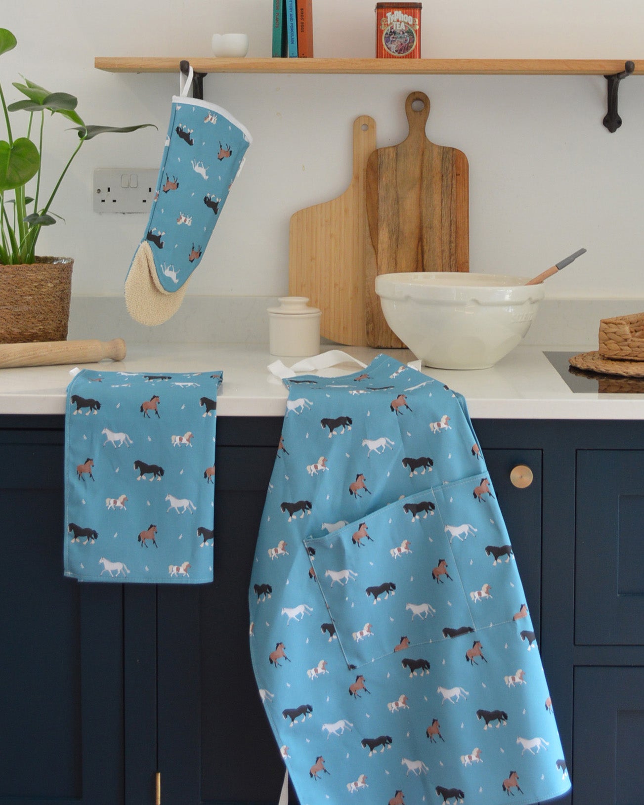Kitchen baking scene with blue apron, oven mitt, and towel featuring horse patterns on a white countertop.