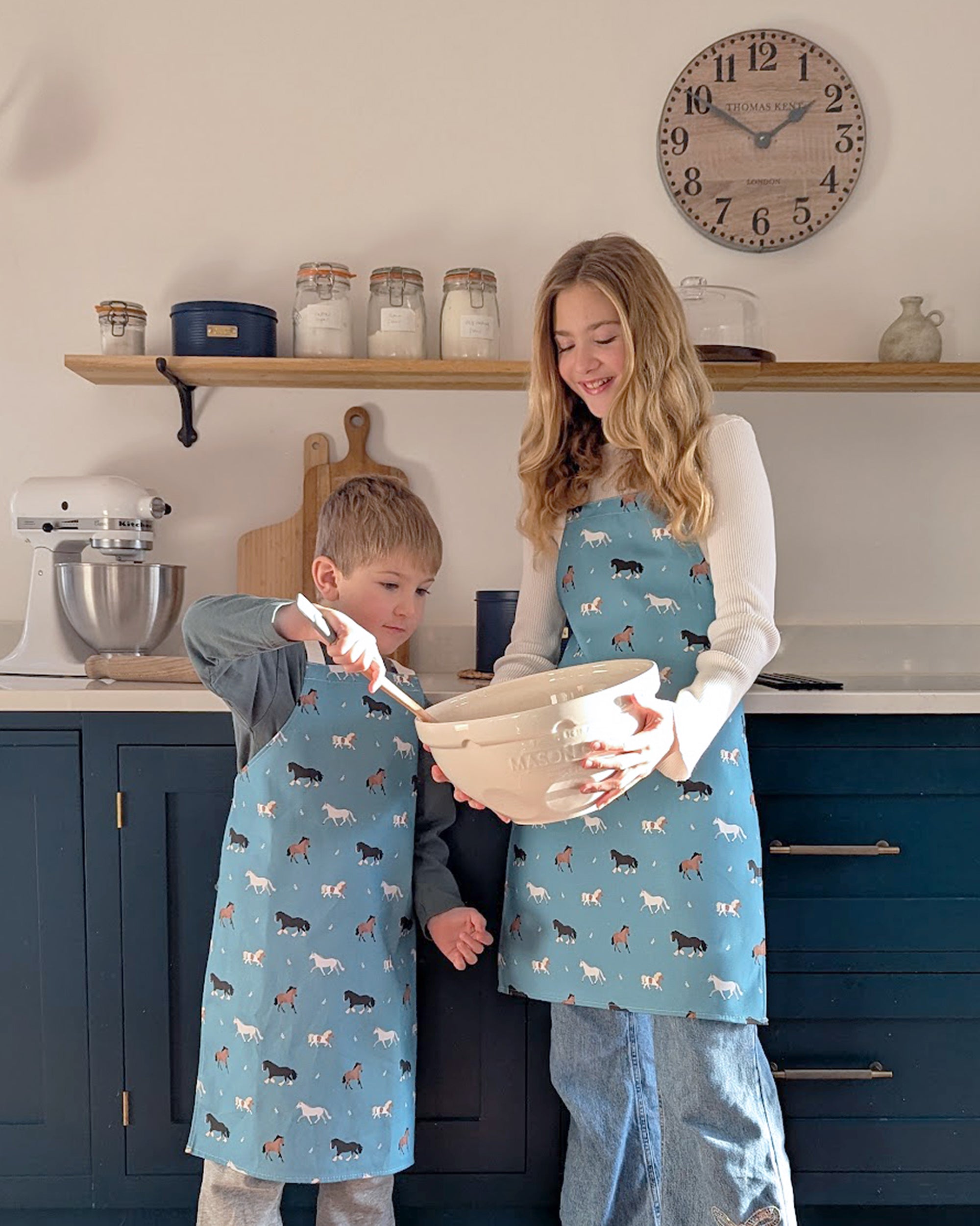 Two children in a kitchen wearing blue aprons with horse and pony patterns, mixing ingredients.