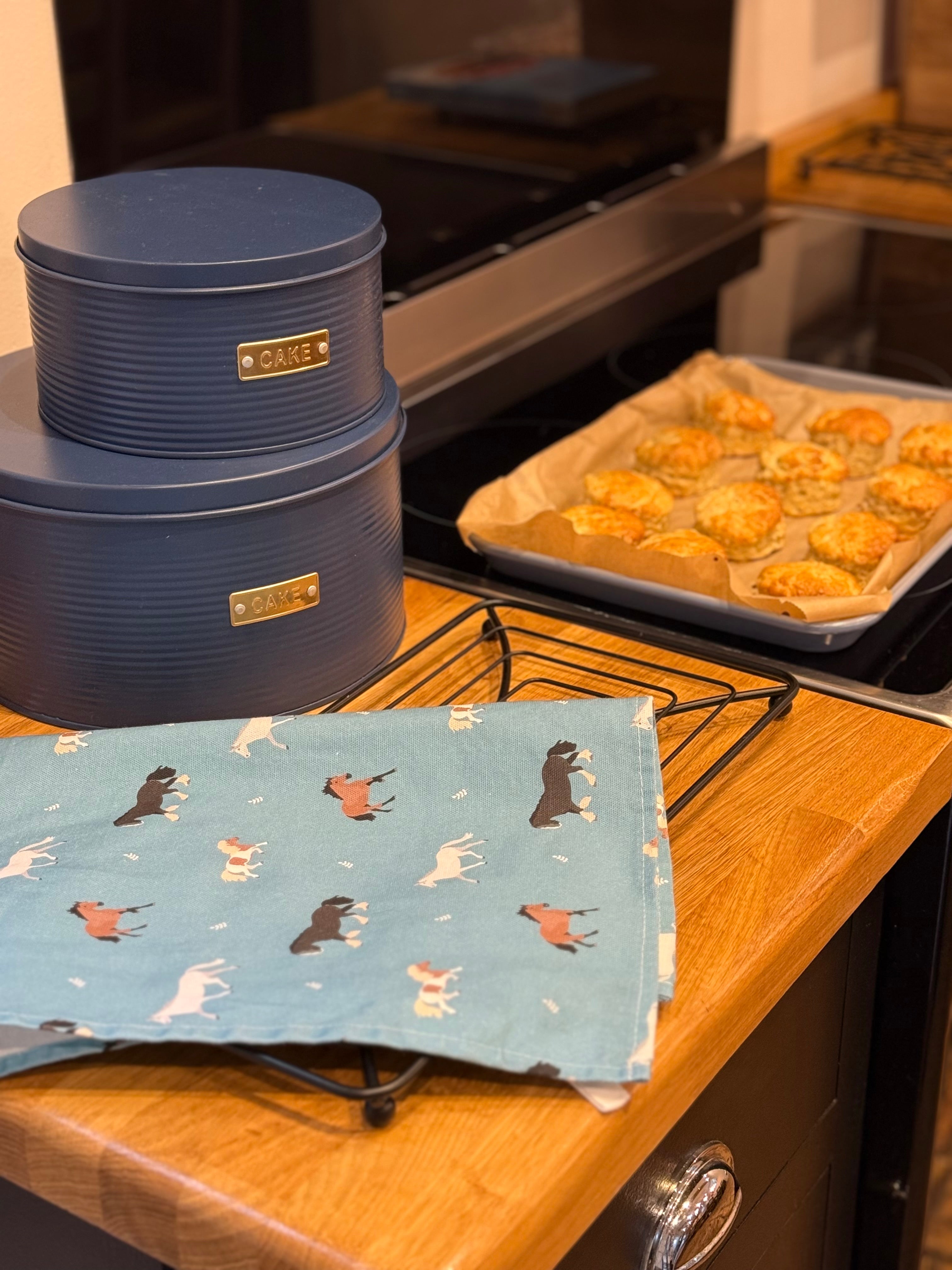Blue horses tea towel on a kitchen worktop with baked scones and cake tins in background