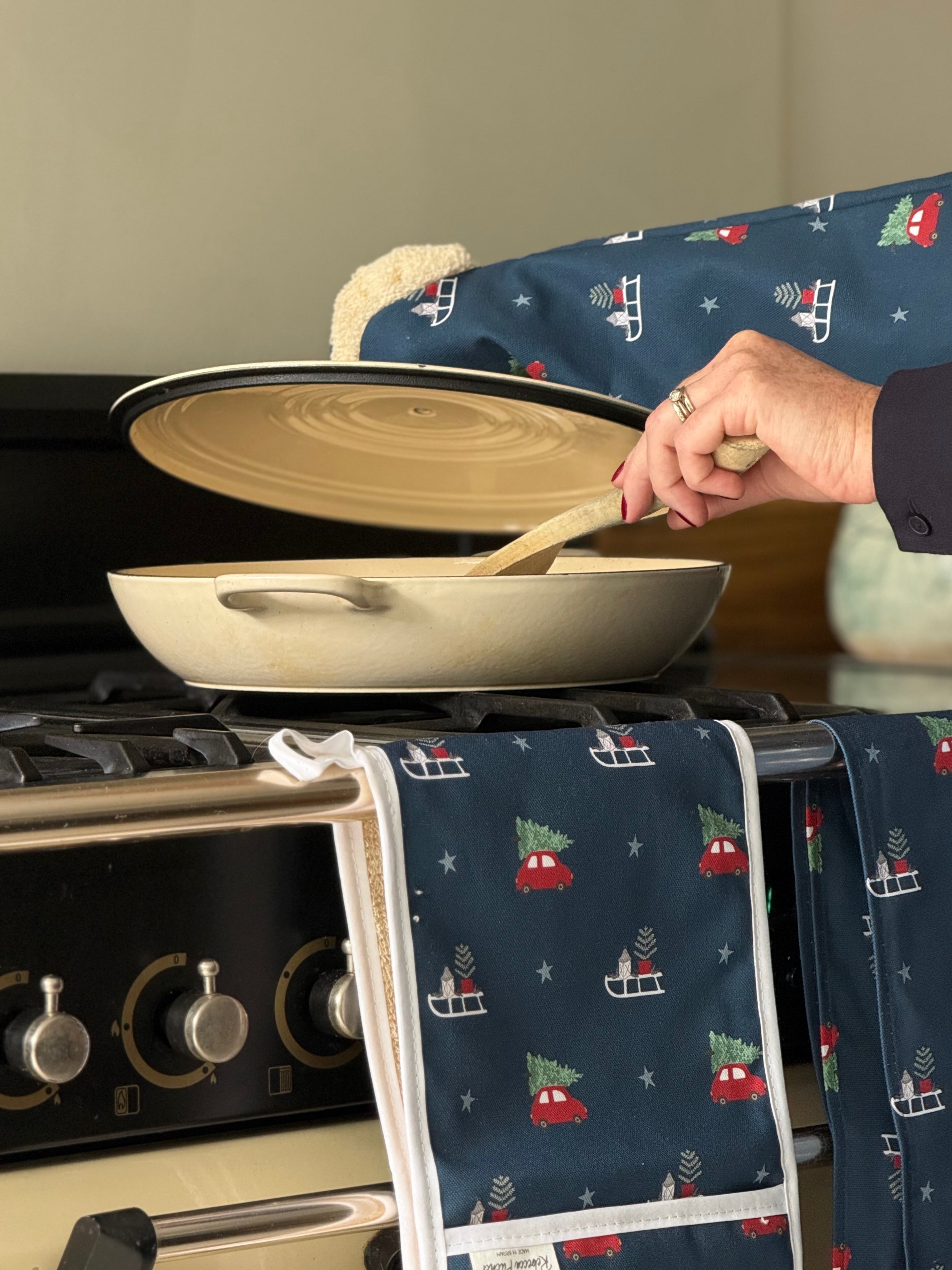 Person using a pan on a stove with Christmas-themed kitchen towels and oven gauntlet hanging nearby.