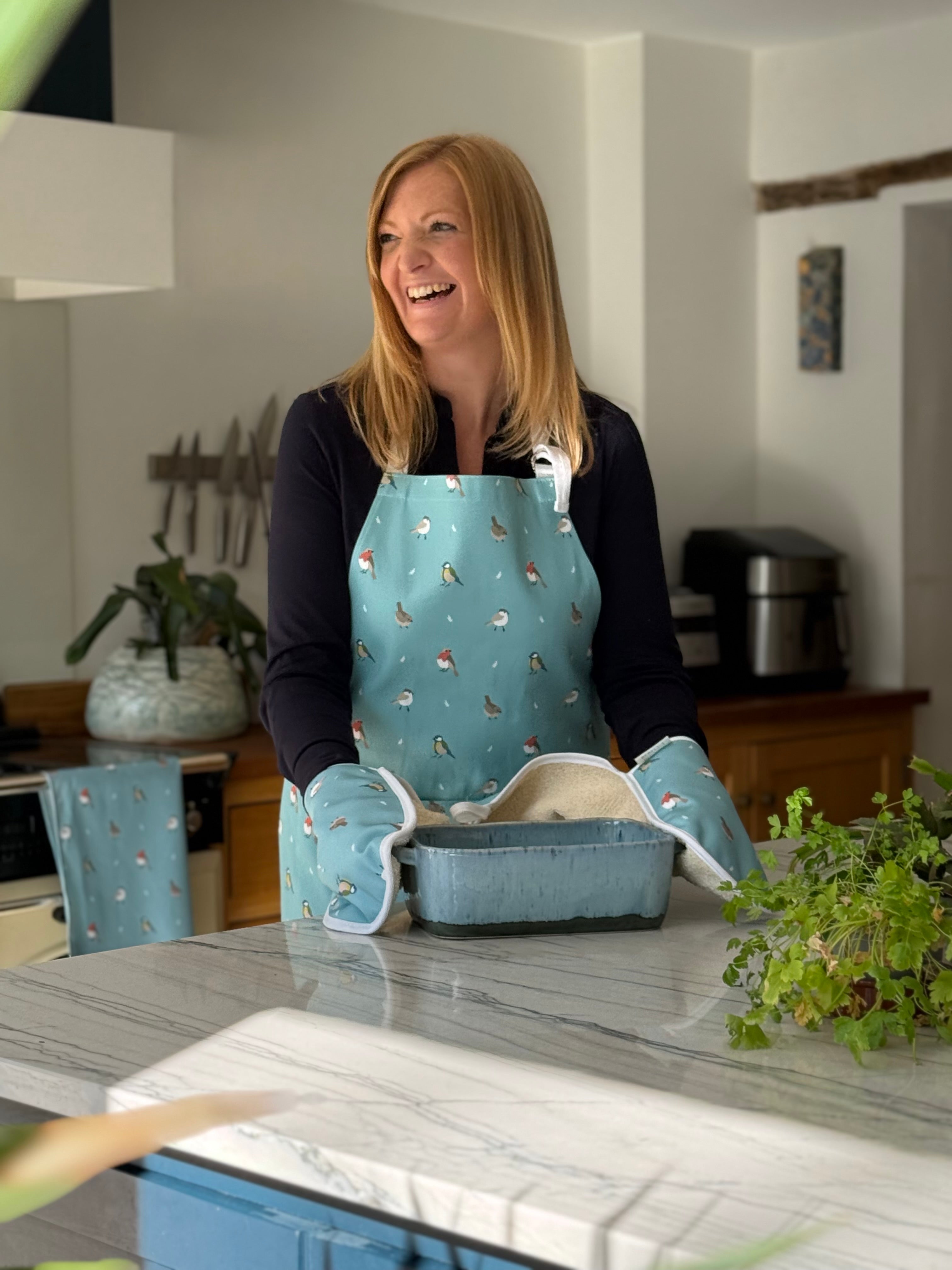 Rebecca Pitcher in a kitchen wearing a blue apron with a bird pattern, standing at a counter with a baking dish.
