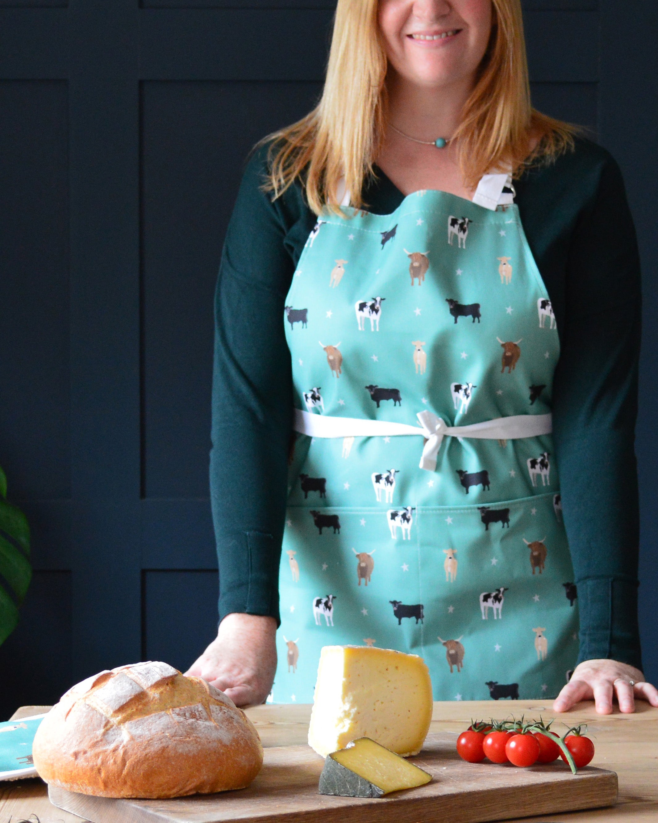 Rebecca Pitcher wearing a green apron with cow pattern standing behind a wooden cutting board with bread, cheese, and tomatoes.