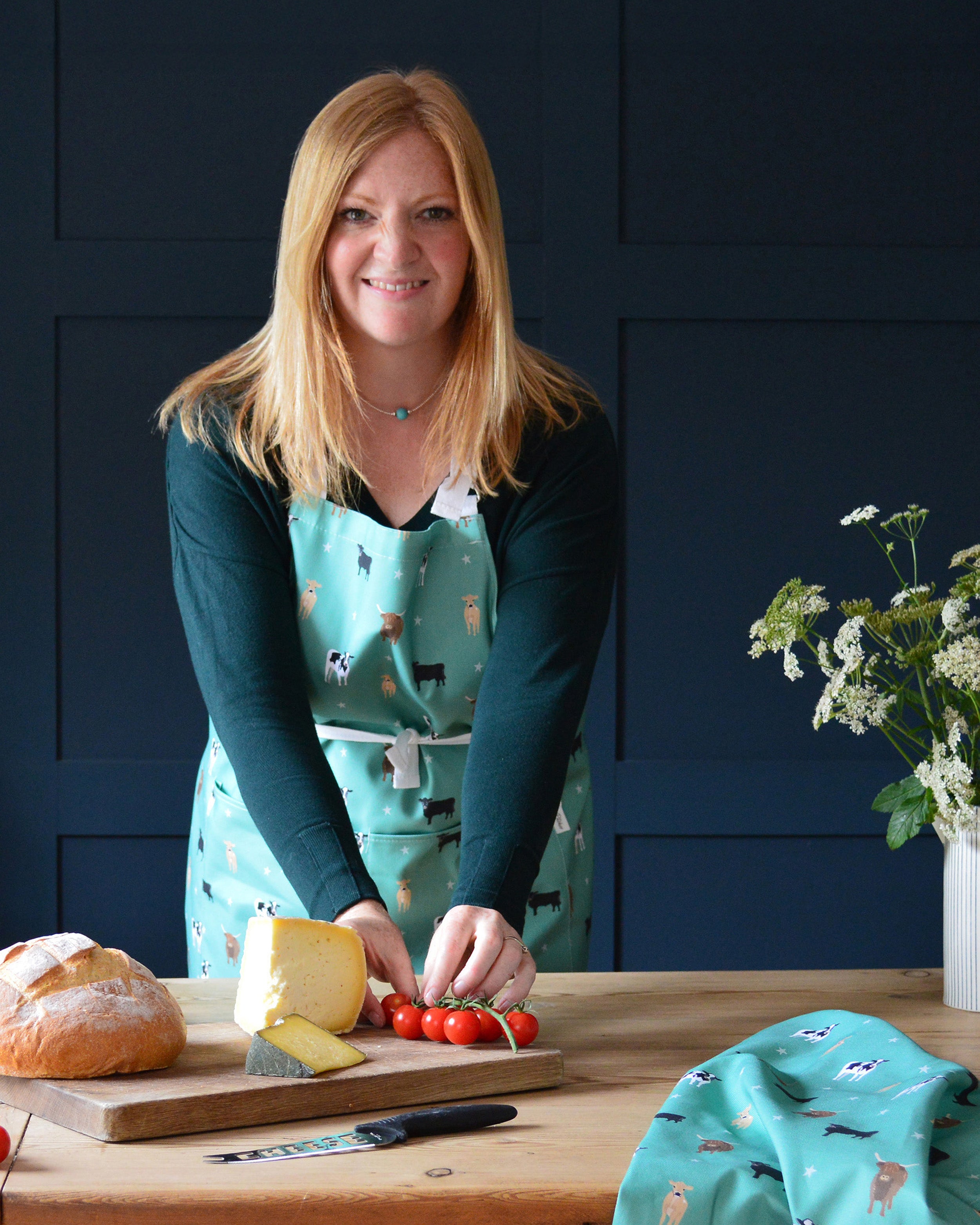 Rebecca Pitcher wearing a green cow apron in a kitchen preparing food with a blue wall background
