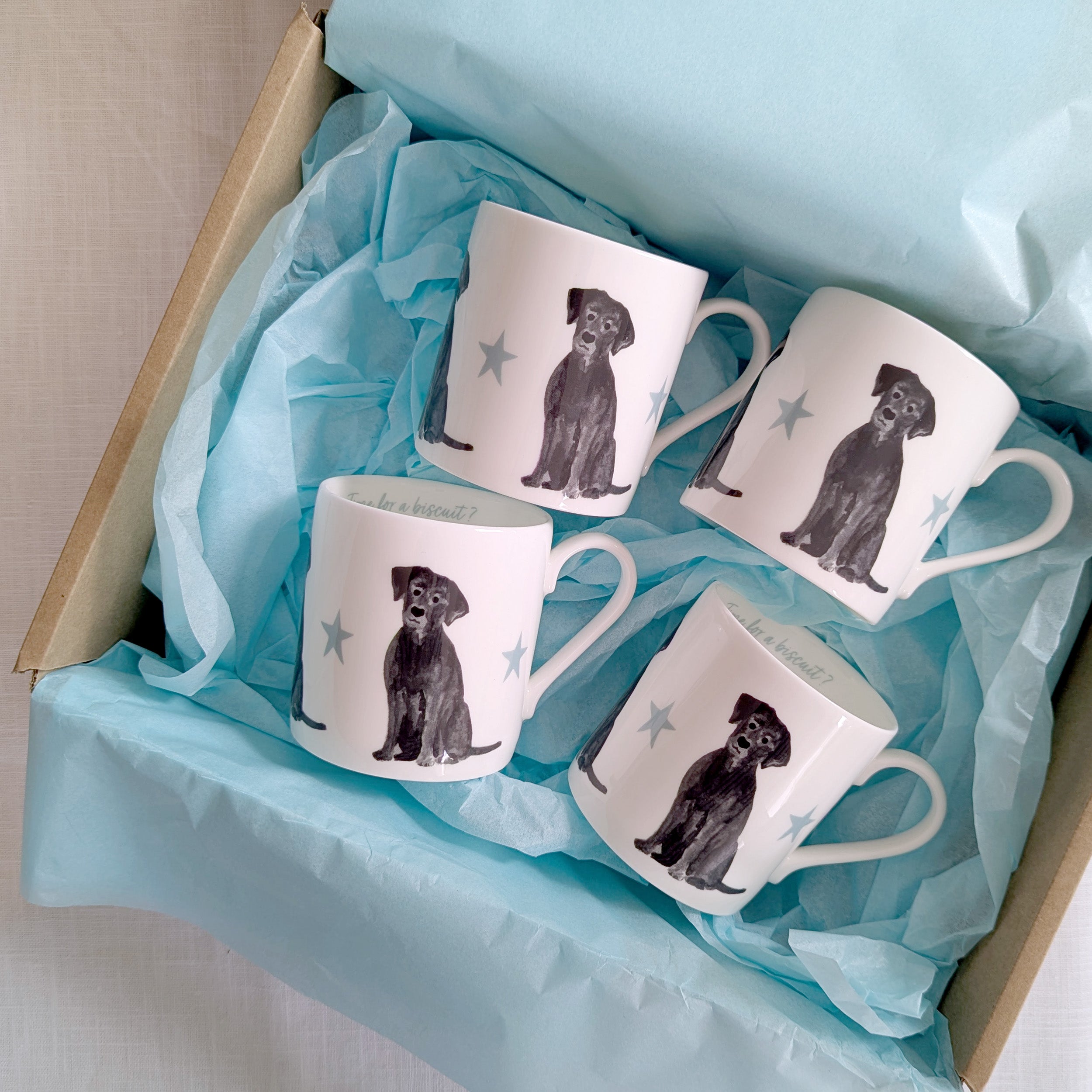 Four white mugs with black labrador dog illustrations in a box lined with blue tissue paper.