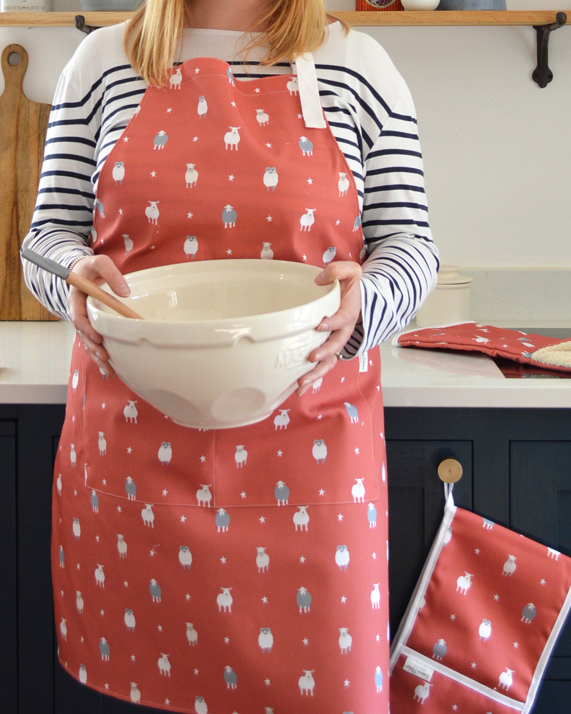 Rebecca Pitcher wearing a red apron with sheep pattern holding a white bowl in a kitchen.