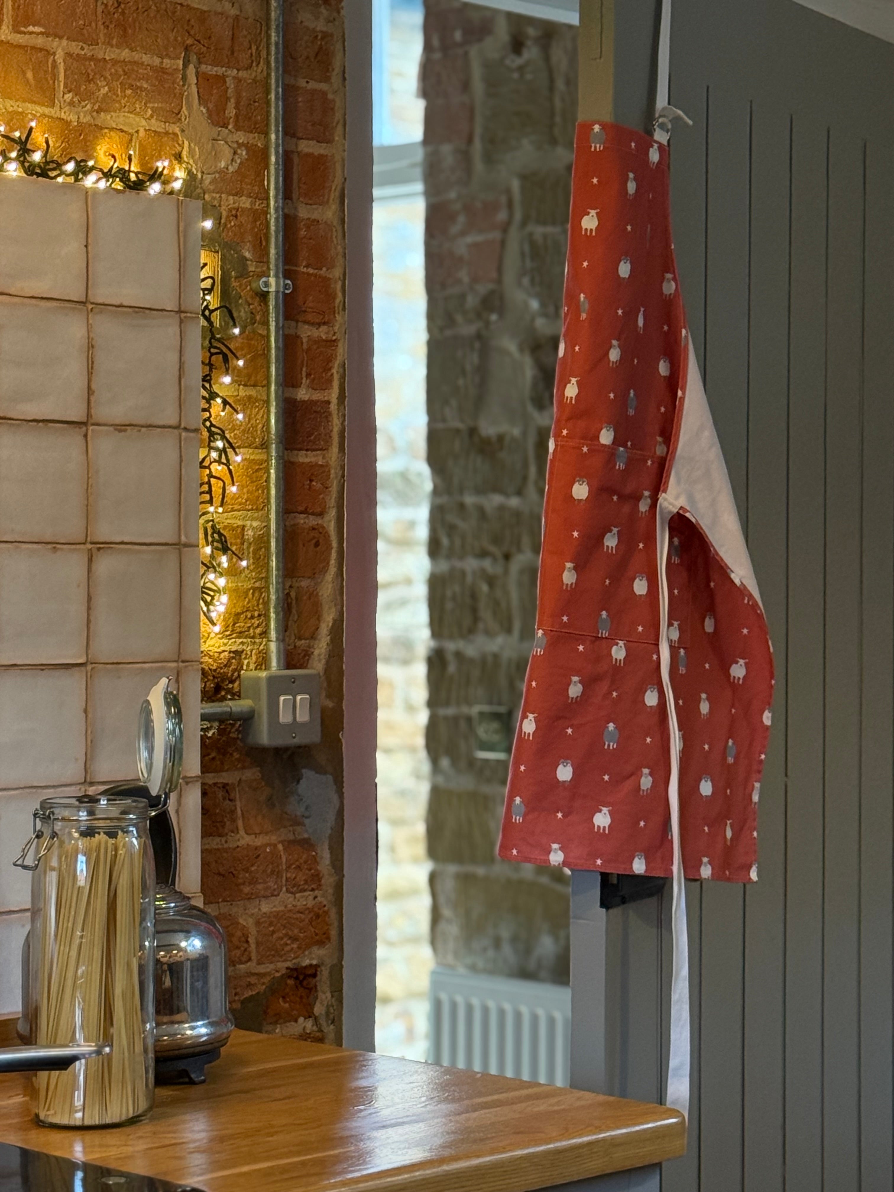 Red apron with white sheep patterns hanging on a wooden door in a kitchen.