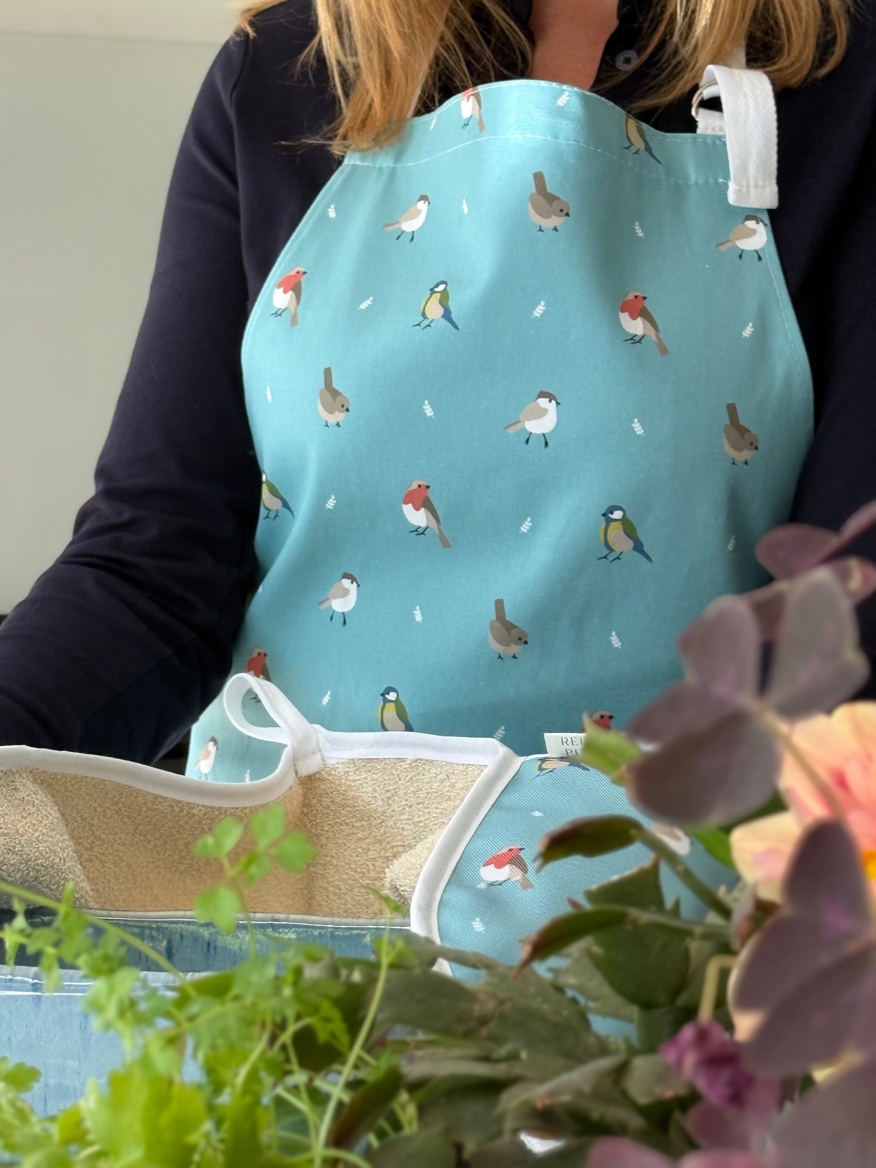Person wearing a blue apron with bird pattern, surrounded by plants and flowers.