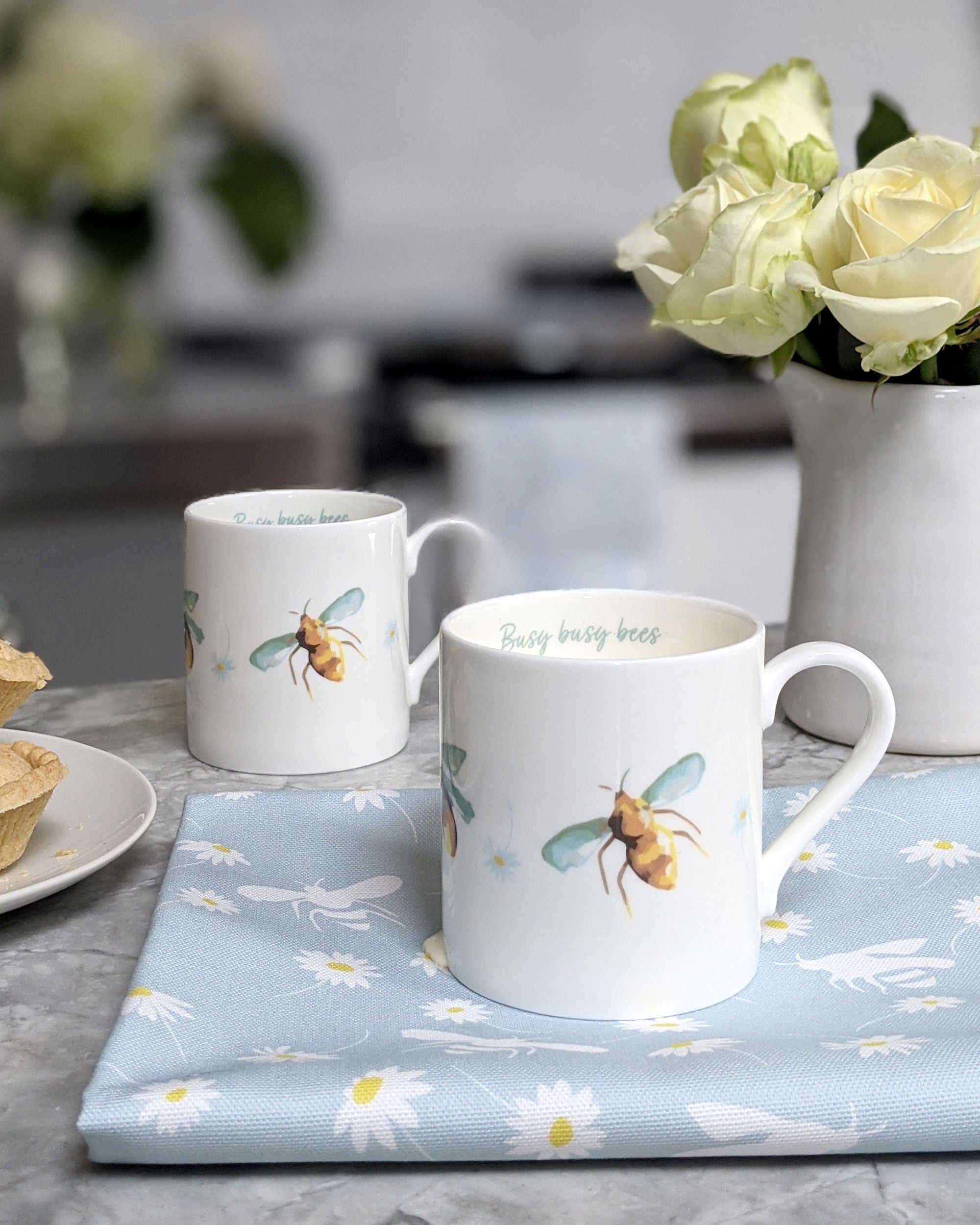 Two mugs with bee designs on a table with a floral arrangement in the background.