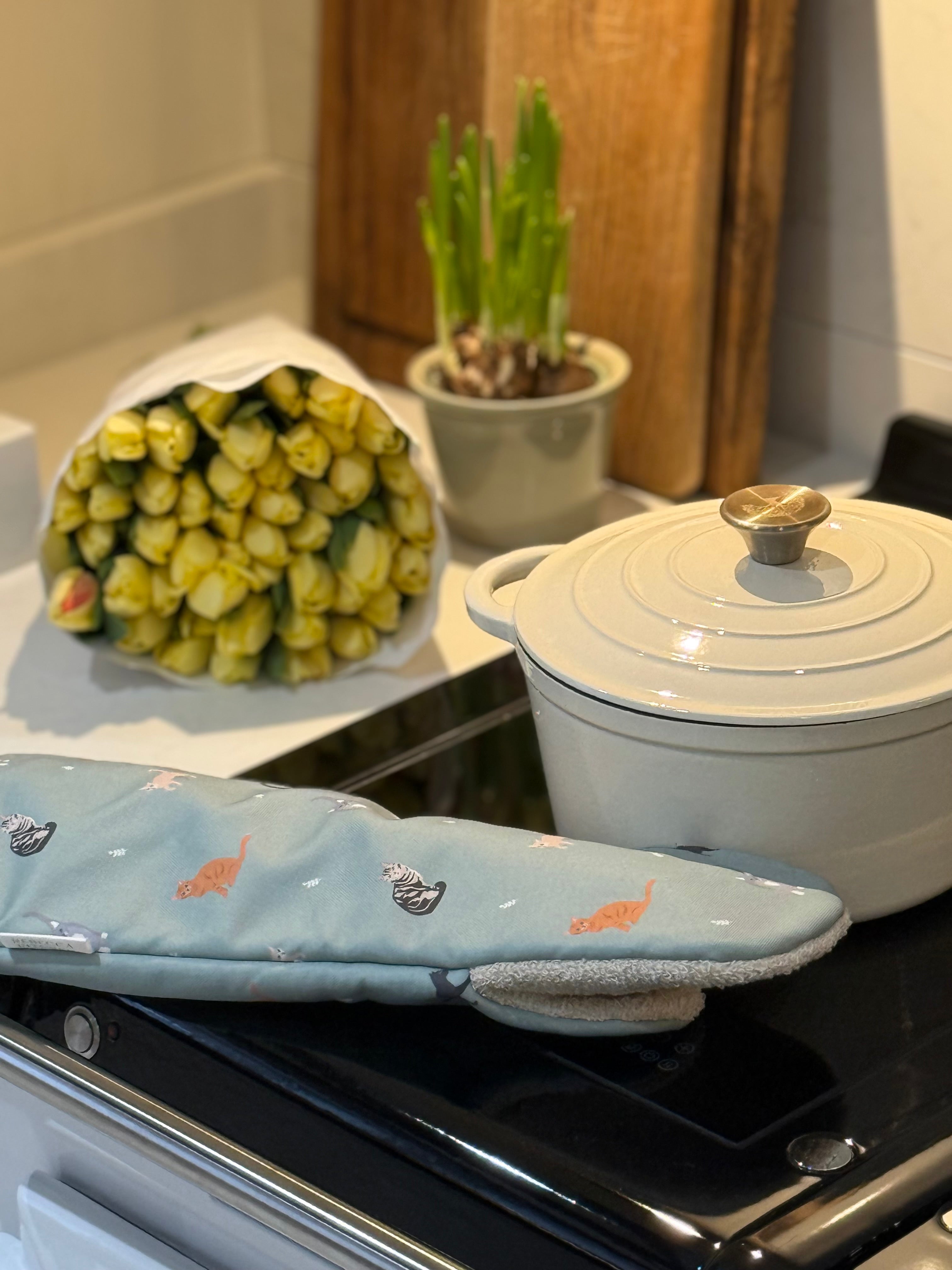 White casserole dish on a stove with a bouquet of yellow flowers and cat patterned oven mit.