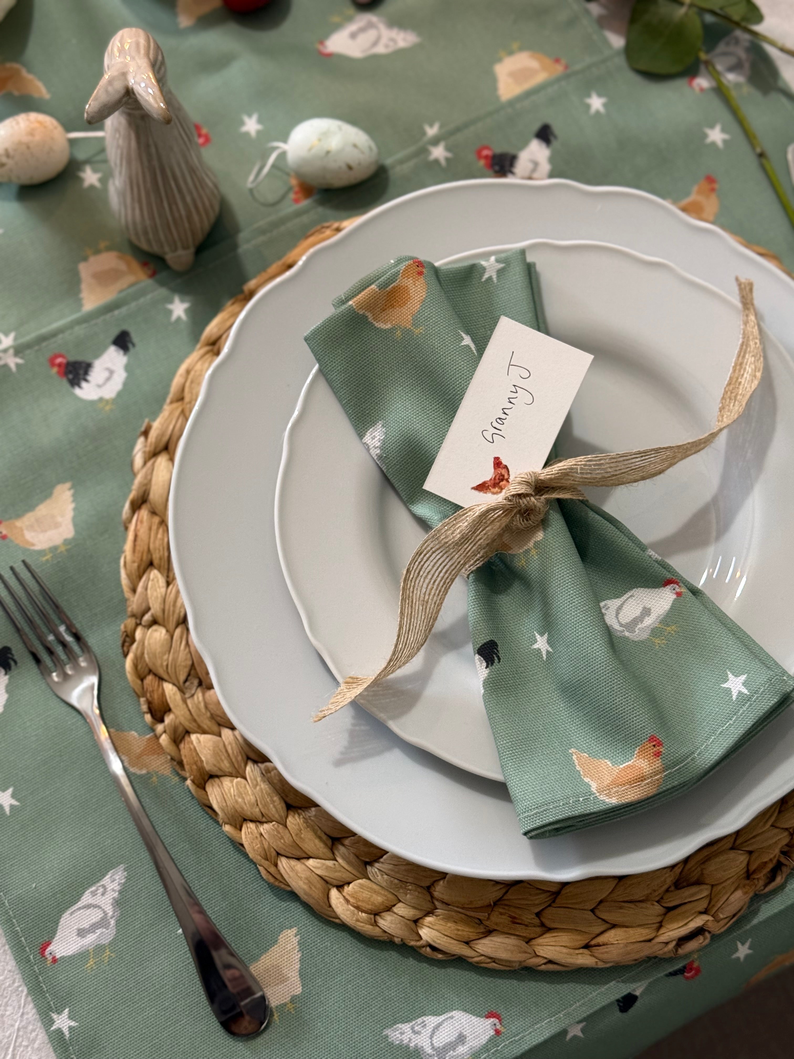 Table setting with a green napkin featuring chicken patterns, white plates, and a woven placemat.