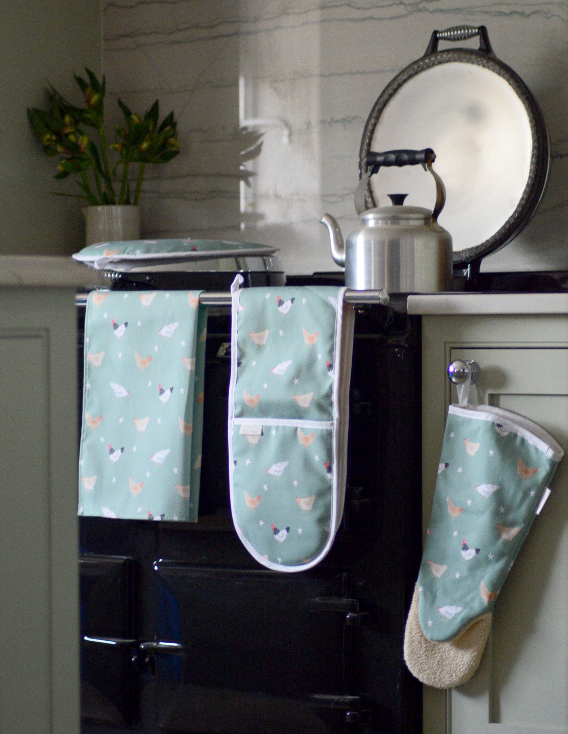 Set of green oven mitts and tea towel with chicken pattern hanging on a stove.