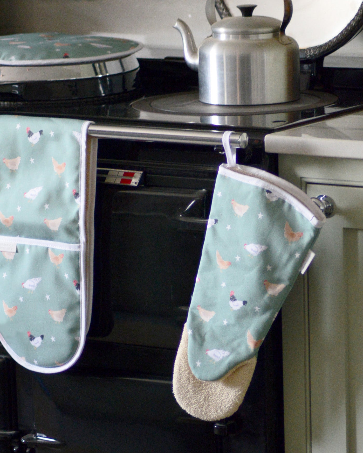Sage green oven mitt with chicken pattern hanging on a stove.