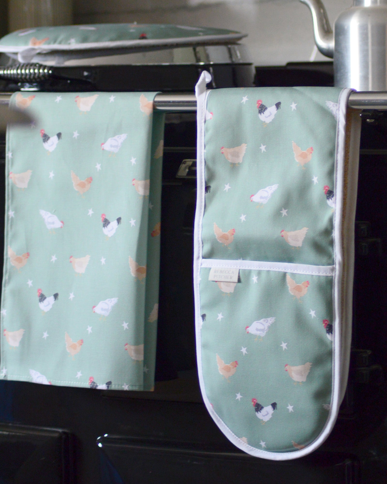 Green oven mitt and tea towel with chicken pattern hanging on a stove.