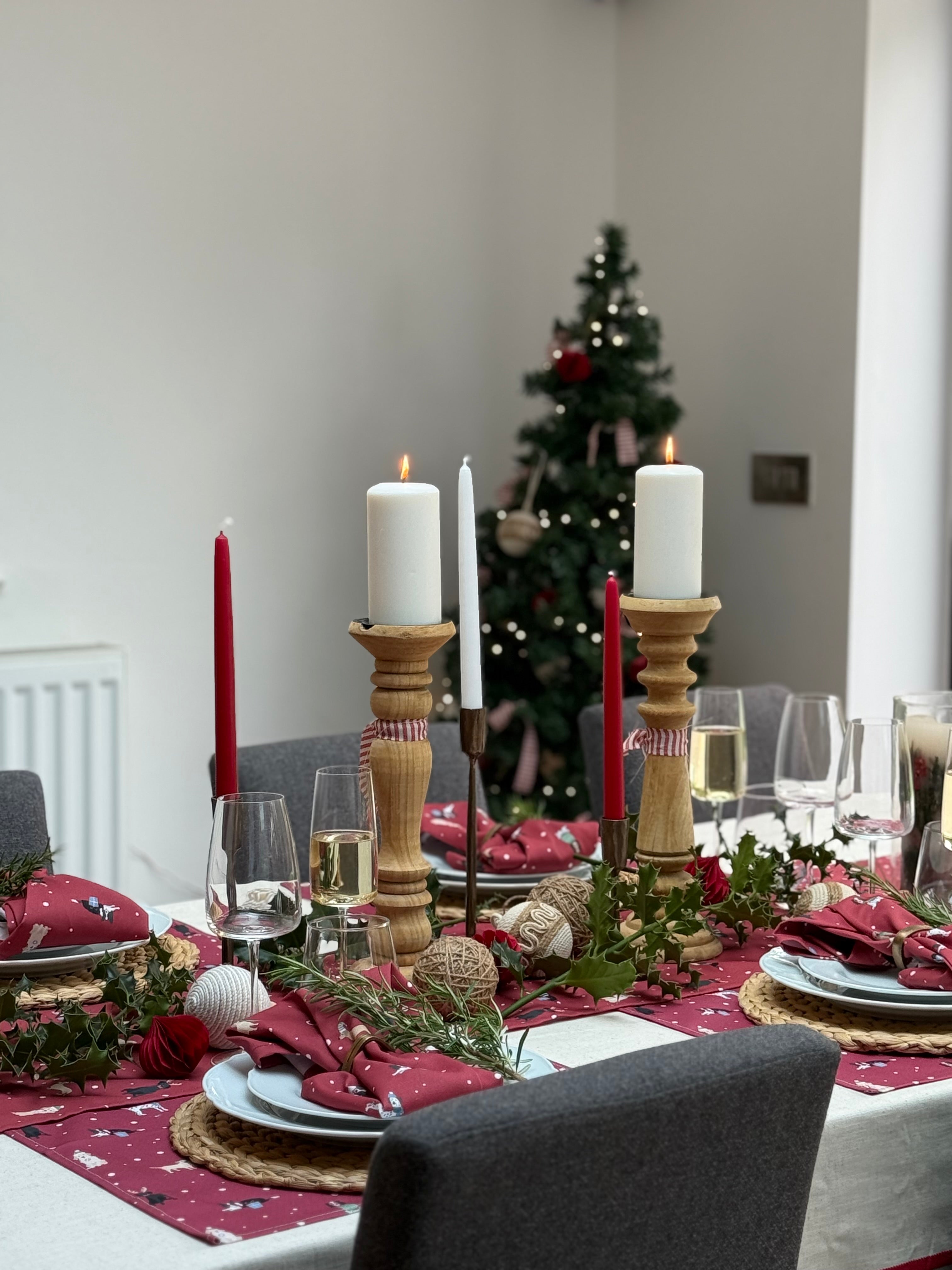 Dining table set for a festive meal with candles, red Christmas dogs runner and napkins, and a Christmas tree in the background.