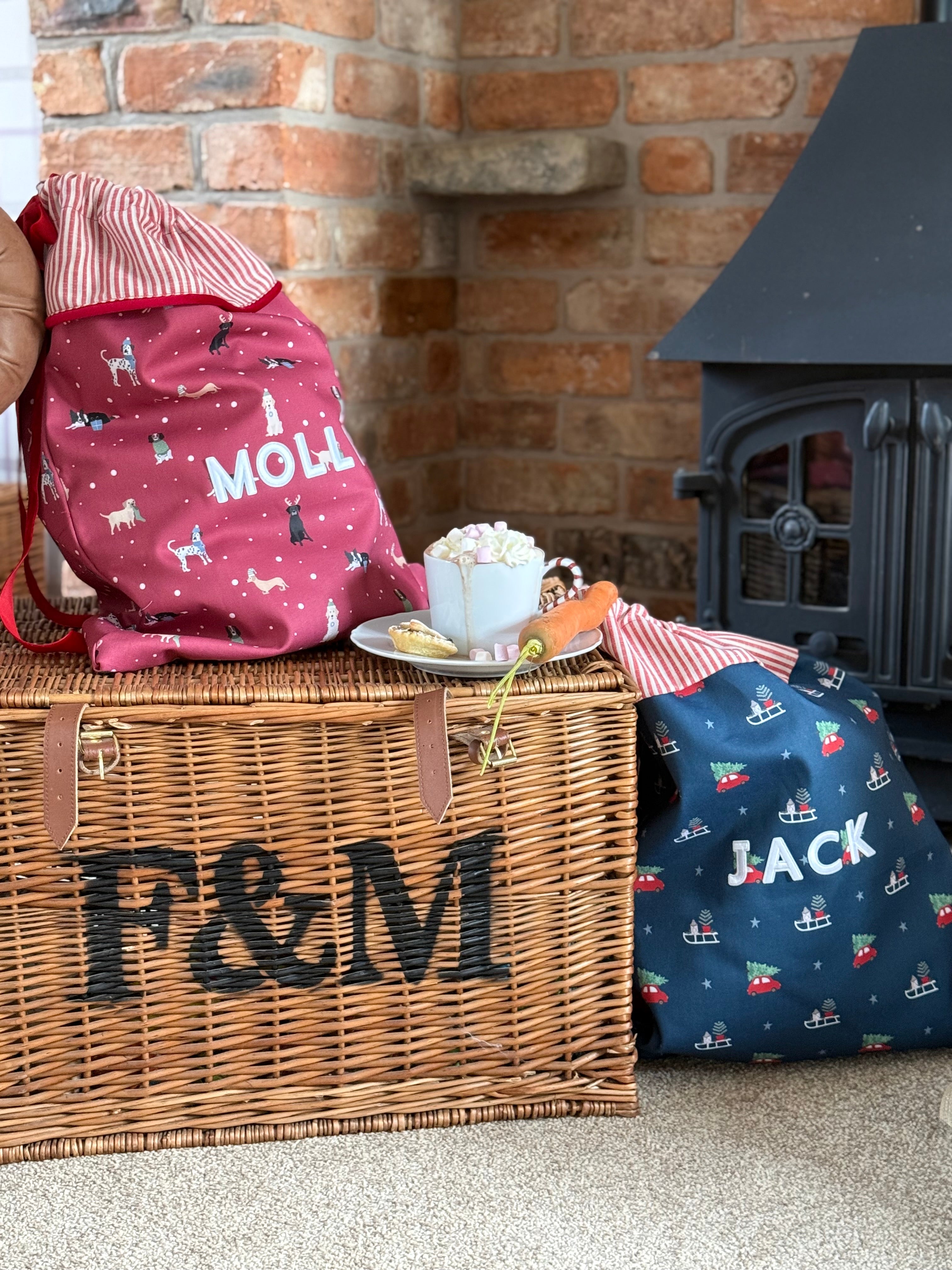 Wicker basket with 'F&M' branding, red santa sack with christmas dog pattern, and navy personalised Christmas sack in front of a brick fireplace.