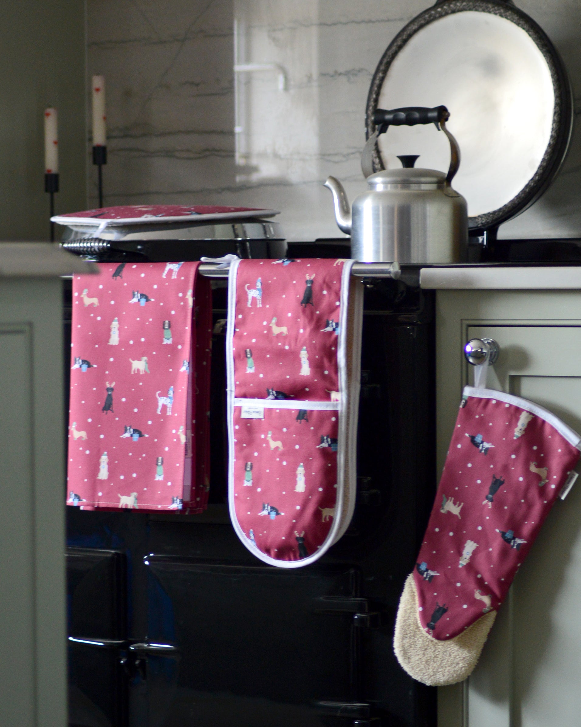 red christmas dogs pattern tea towel, oven mitt, and hob cover hanging on a black range cooker in a kitchen.