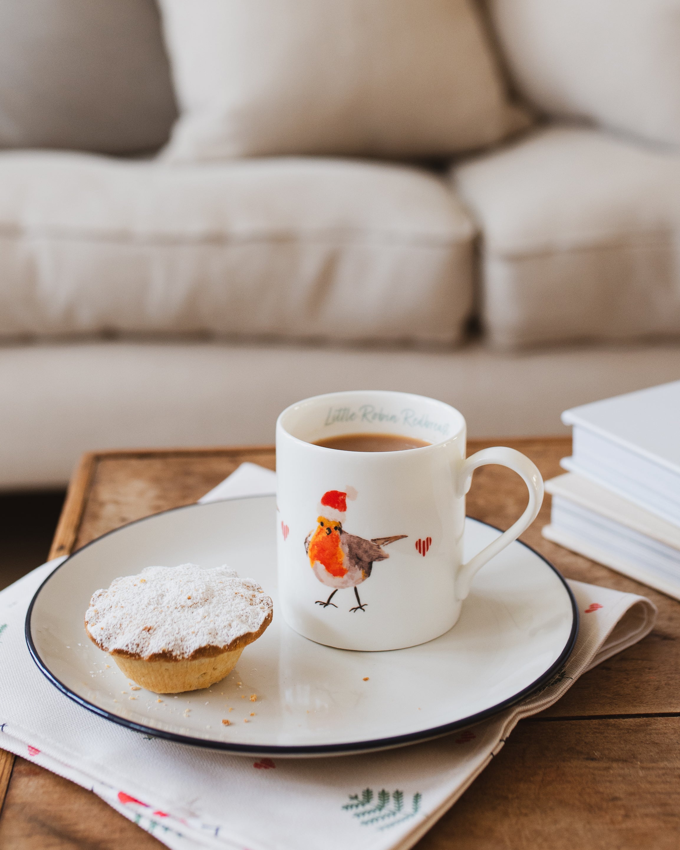 Coffee mug with a robin design on a saucer, next to a mince pie on a wooden table.